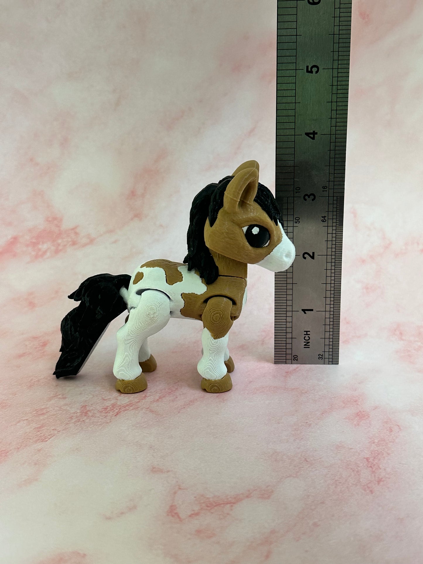 3d printed toy brown and white horse figure with a ruler for scale on a pink background