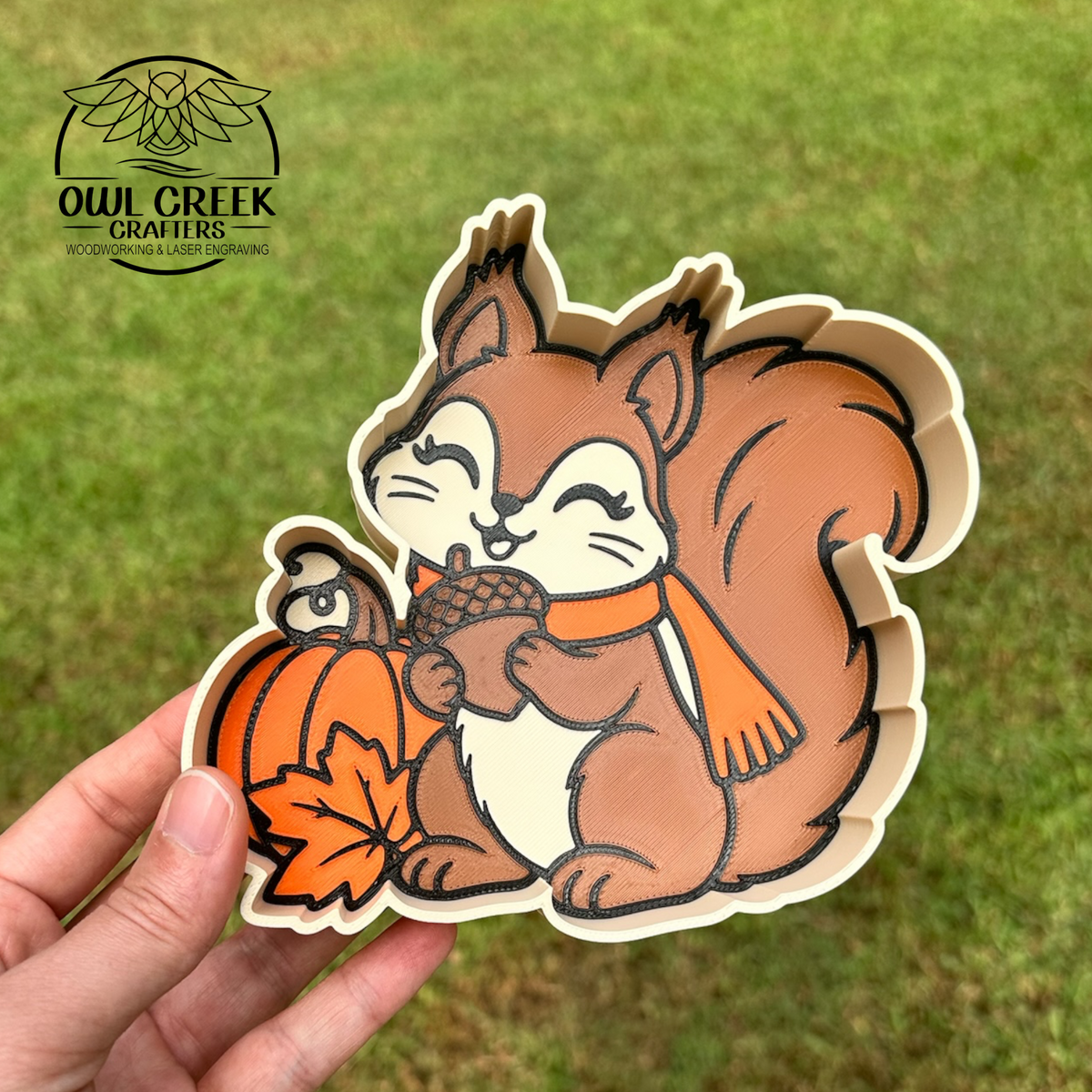 A hand holds a 3d printed squirrel shaped trinket tray decorated with a brown squirrel holding an acorn with an orange pumpkin and orange leaf next to it, set against a green grass background. The Owl Creek Crafters logo is visible in the top left corner.
