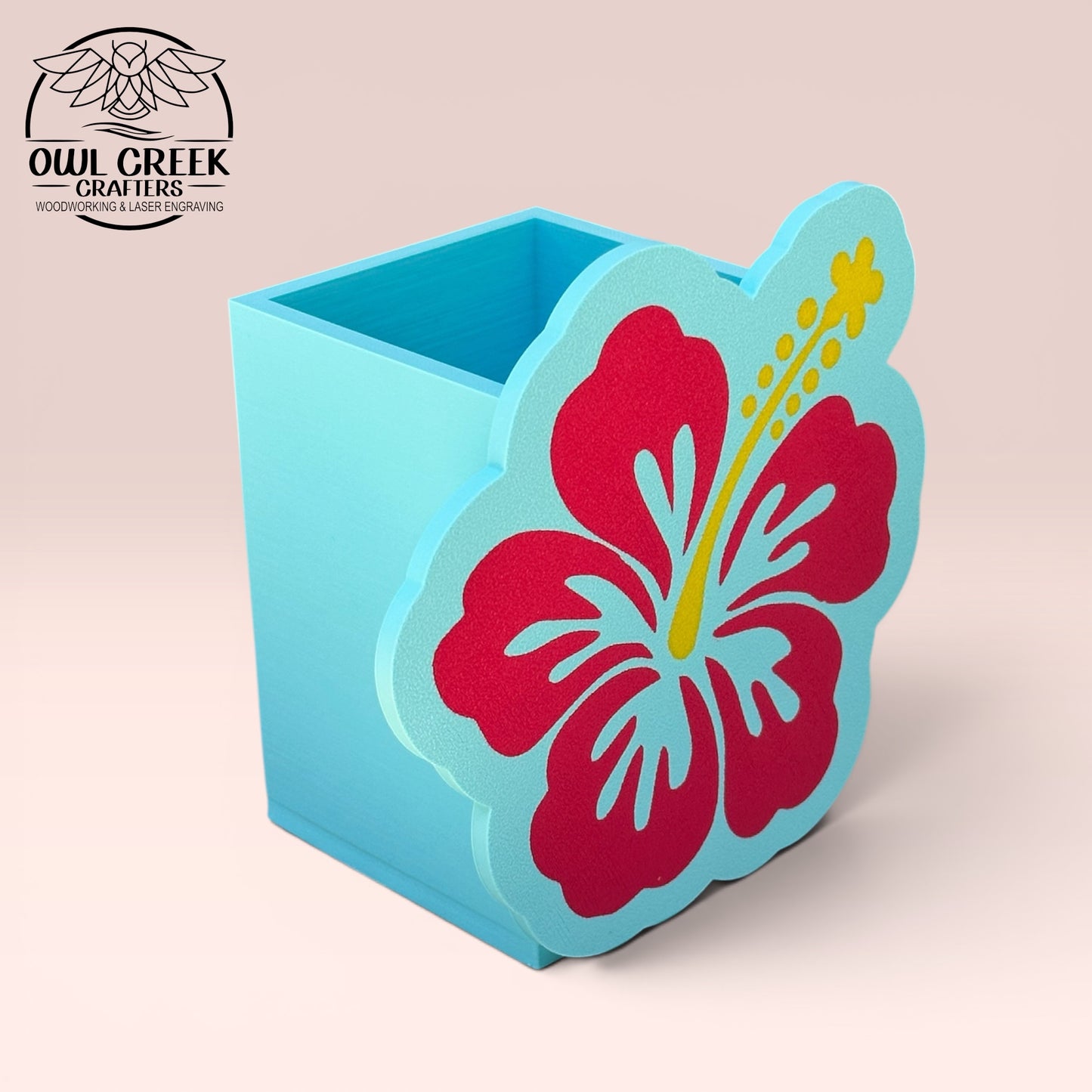 A decorative 3d printed hibiscus flower pencil holder in magenta and light blue with a yellow stamen, displayed on a light pink background. The Owl Creek Crafters logo is visible in the top left corner.