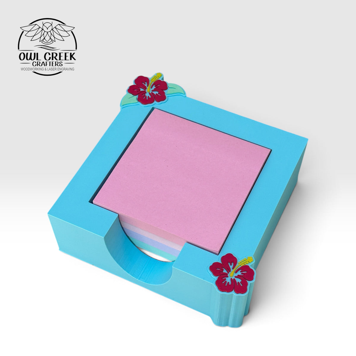 A 3d printed light blue sticky note holder with two magenta hibiscus flowers in the top left and bottom right corners. On a light beige background with the Owl Creek Crafters logo in the top right corner.