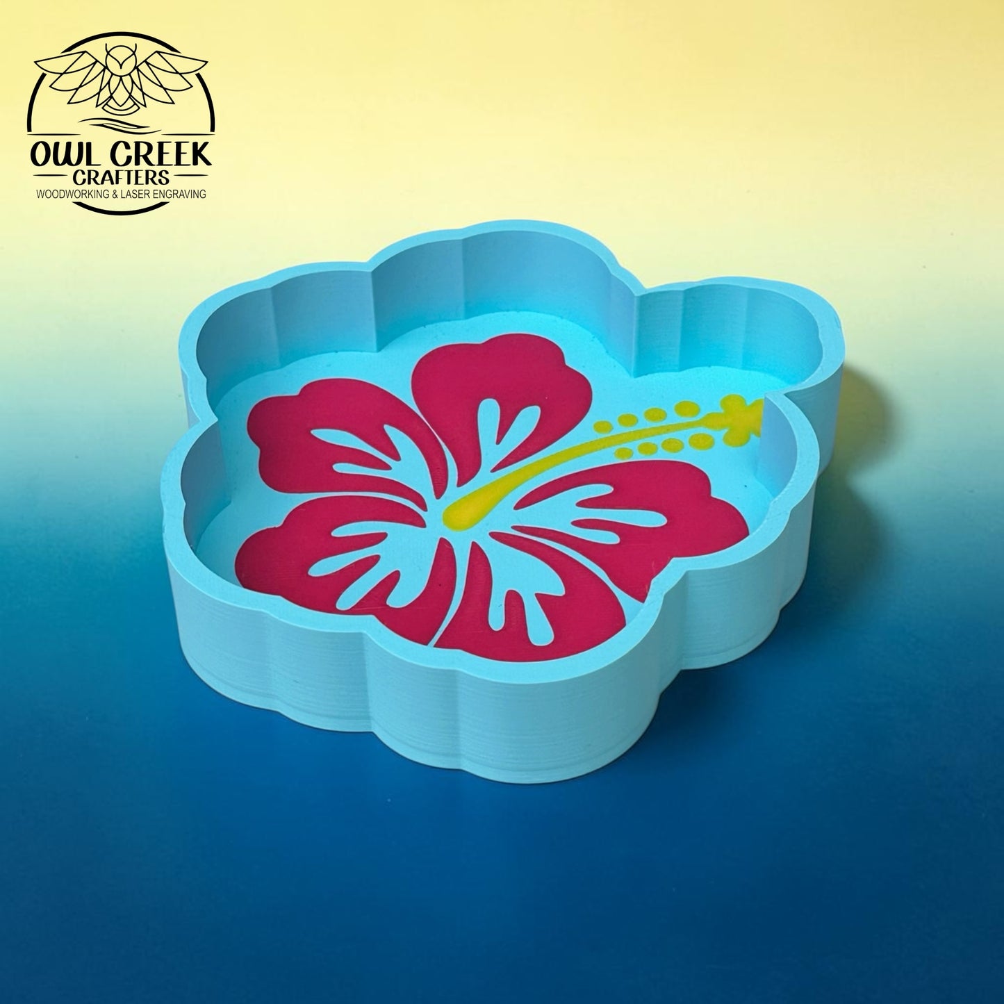 A light blue flower-shaped trinket tray with a pink hibiscus flower and yellow stamen design printed inside, shown on a blue-to-yellow gradient background. The Owl Creek Crafters logo is visible in the top-left corner.