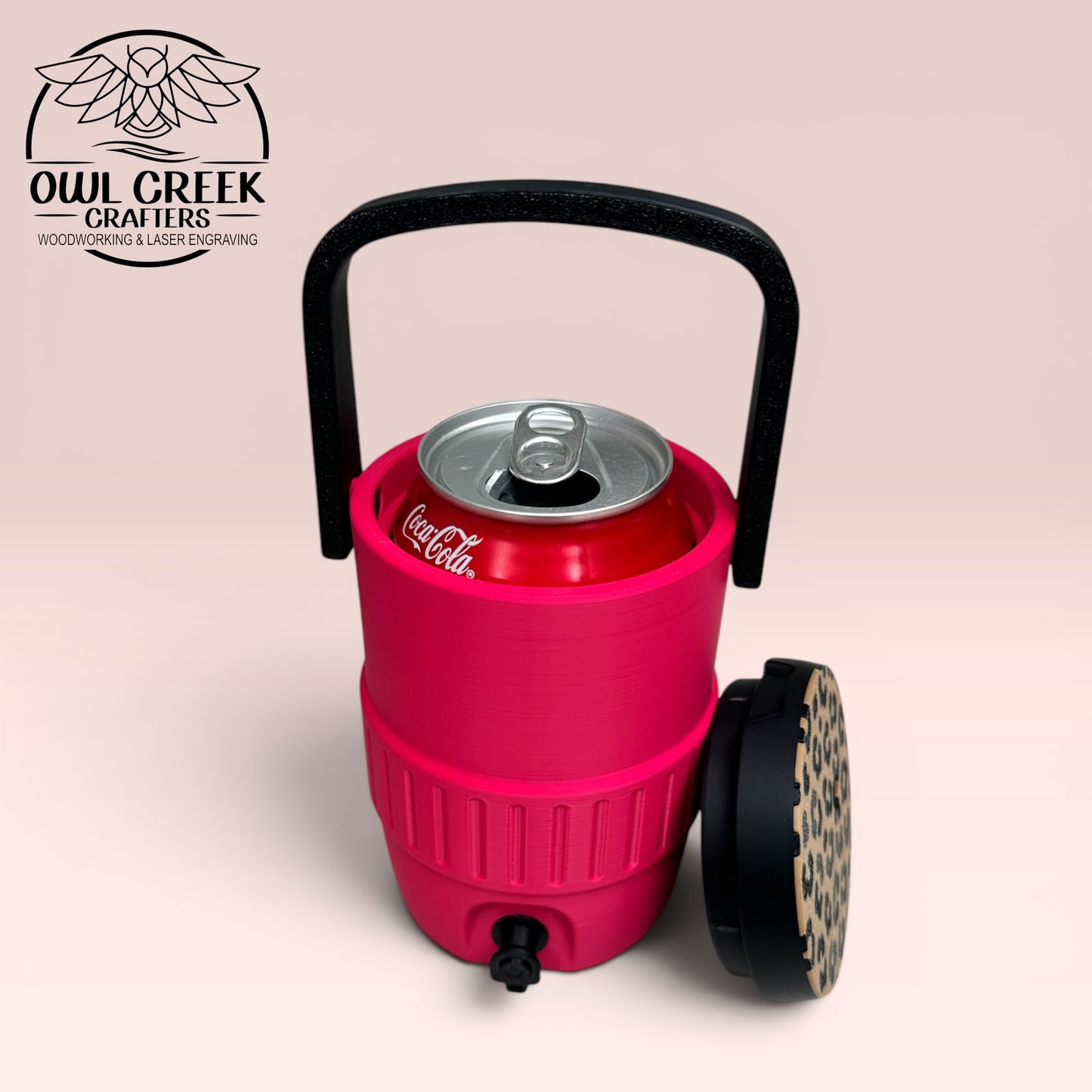 Pink can cooler coozie with a black handle, a Coca-Cola can inside it, and a leopard print lid leaning against the side, on a beige background with the Owl Creek Crafters Logo