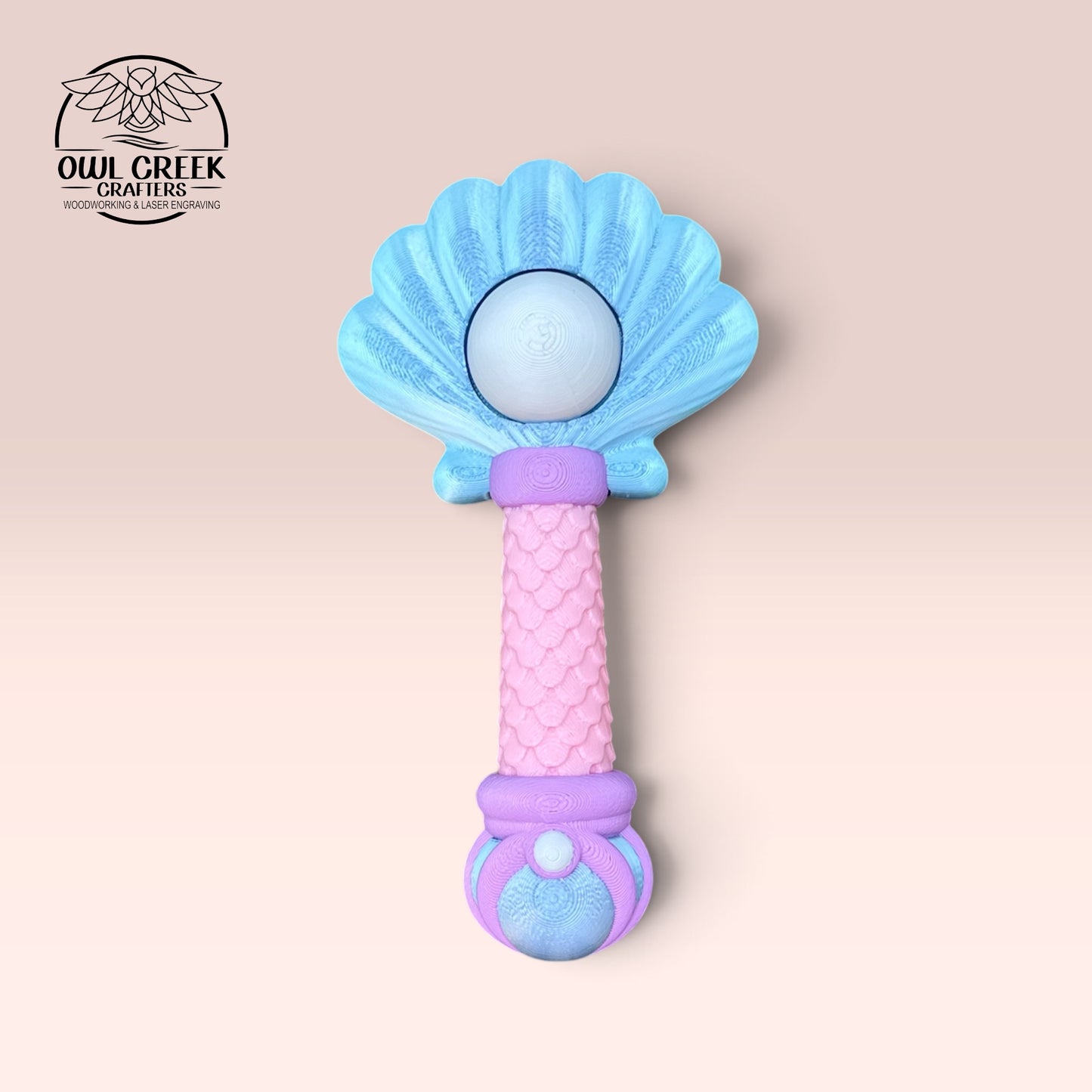 A 3d mermaid clicker wand, featuring a blue shell-shaped top, a pink textured handle, and purple and pink accents, shown on a light pink background with the Owl Creek Crafts logo in the top left corner.