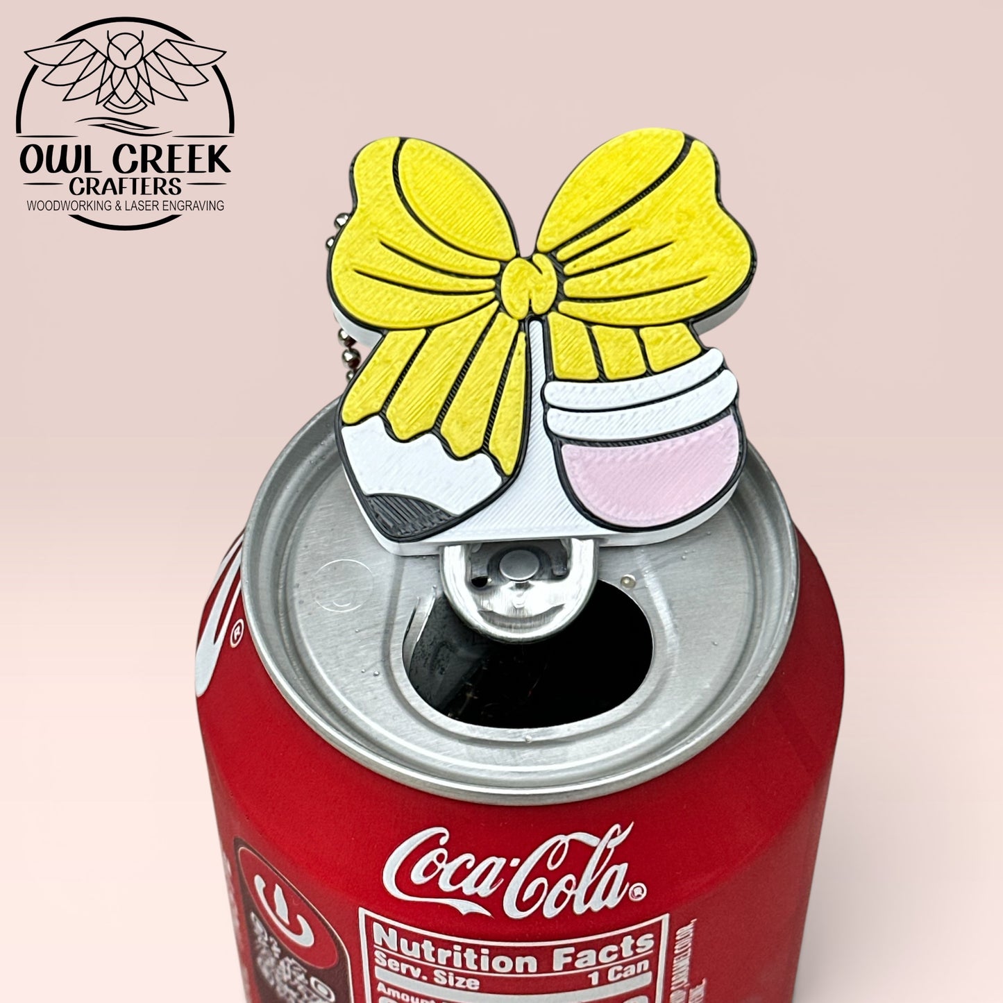 Bow in the shape and color of a pencil design keychain on the tab of a Coca-Cola can, with a beige background and the Owl Creek Crafters Logo