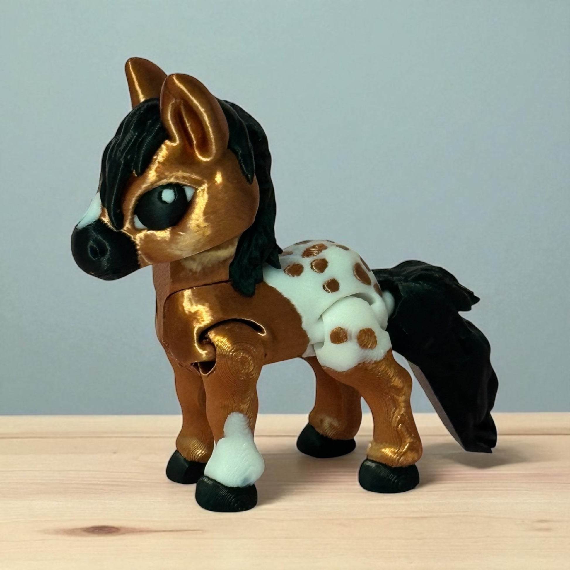 3d printed toy horse with brown body, black mane and tail, and white spots on a wooden surface and light gray background