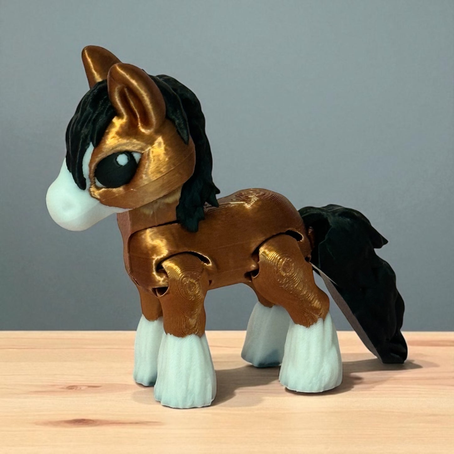 3d printed toy horse figure with brown body, black mane and tail, and white feet on a wooden surface with a gray background.