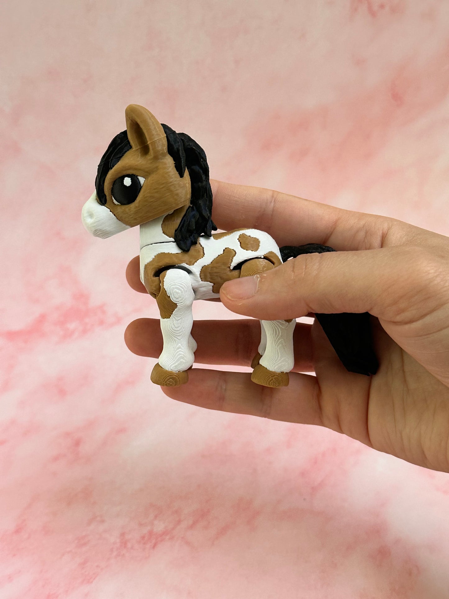 Hand holding a 3D printed brown and white horse toy against a pink background