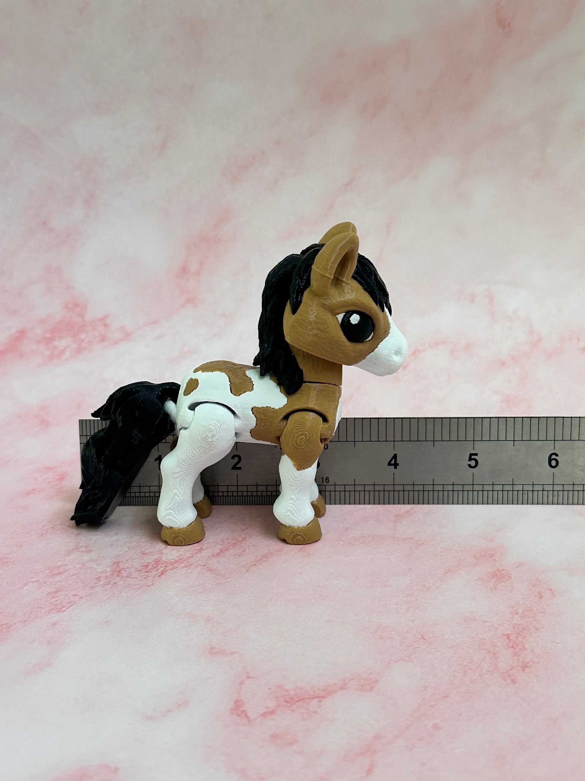 3d printed toy brown and white horse figure next to a ruler on a pink background