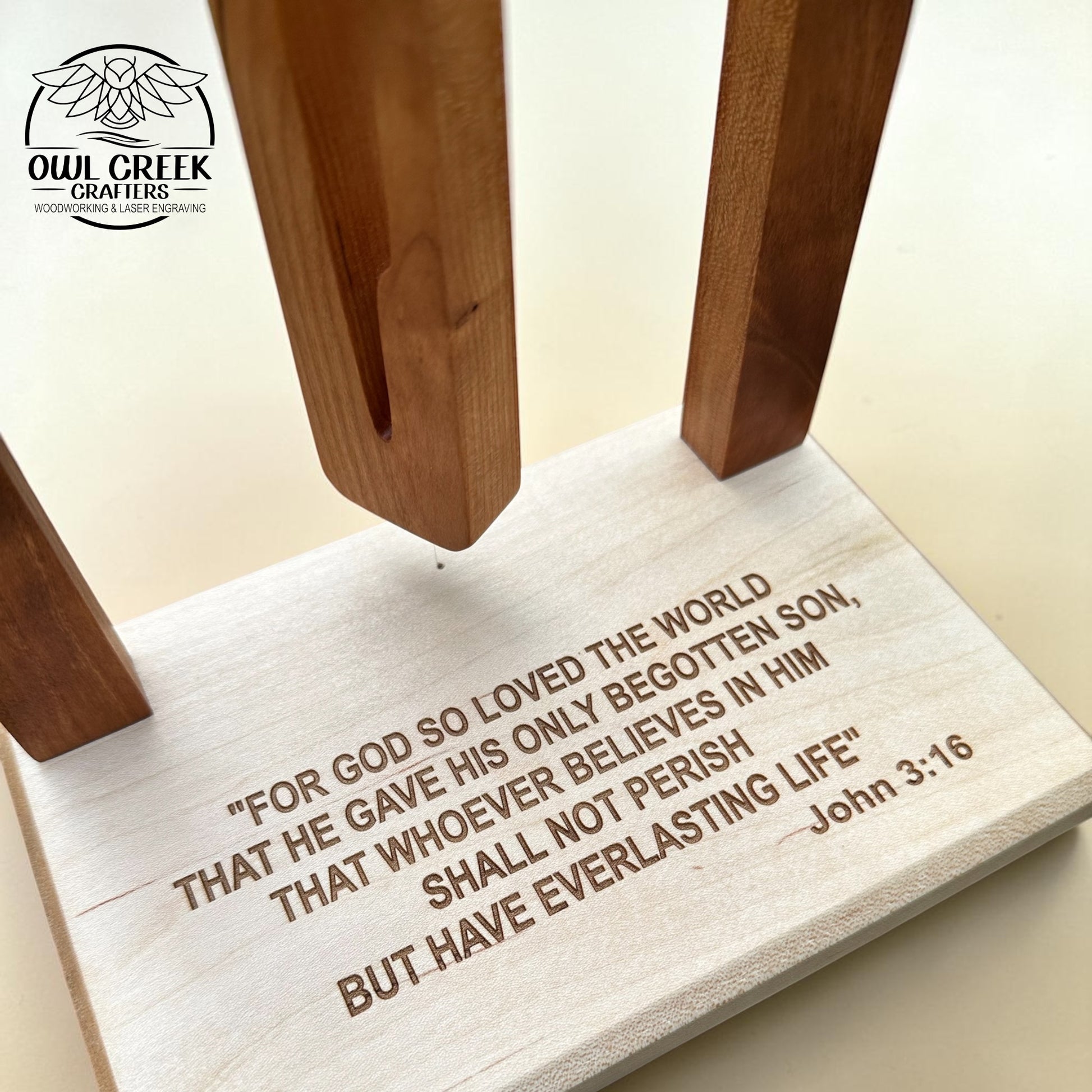 A wooden Bible verse plaque or stand with engraved text reading John 3:16, shown on a light beige background. The base is pale natural wood with dark brown engraved lettering, and several upright wooden supports extend upward.