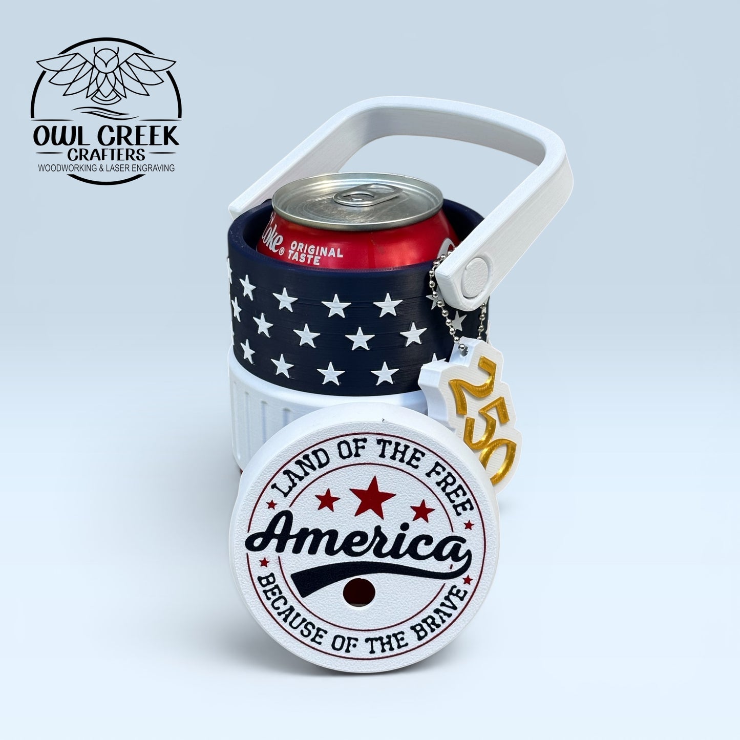 American flag-themed can cooler coozie and a '250' keychain, a coke can inside the coozie, with a red white and blue lid that says 'America land of the free because of the brave', on a light gray background with the Owl Creek Crafters logo