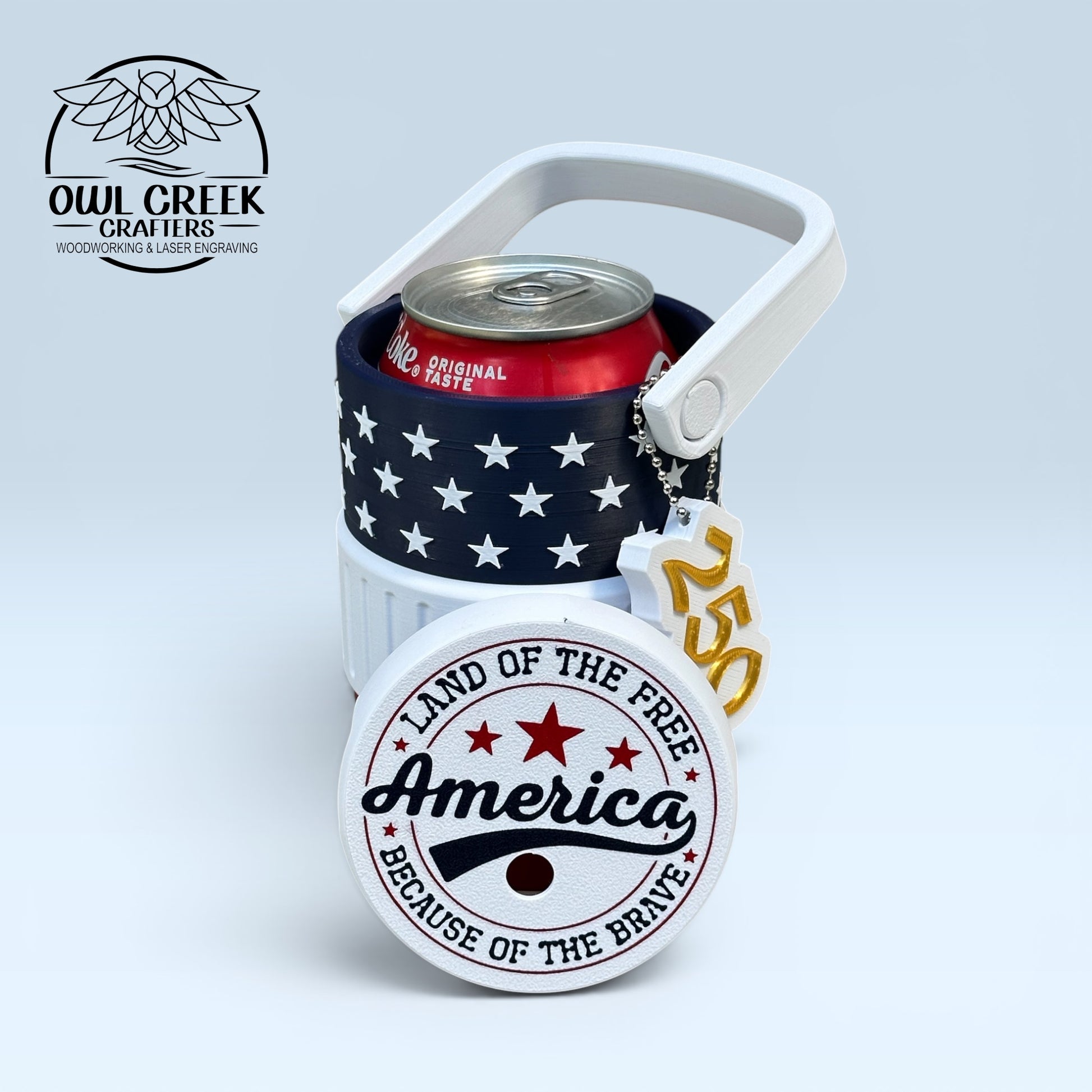 American flag-themed can cooler coozie and a '250' keychain, a coke can inside the coozie, with a red white and blue lid that says 'America land of the free because of the brave', on a light gray background with the Owl Creek Crafters logo
