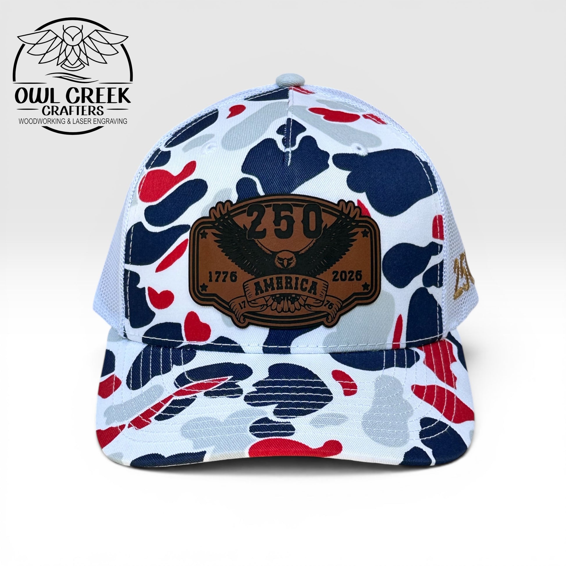 Red, white, and blue camouflage pattern hat with a leatherette patch that says "250 America, 1776-2026", with a light beige background and the Owl Creek Crafters Logo