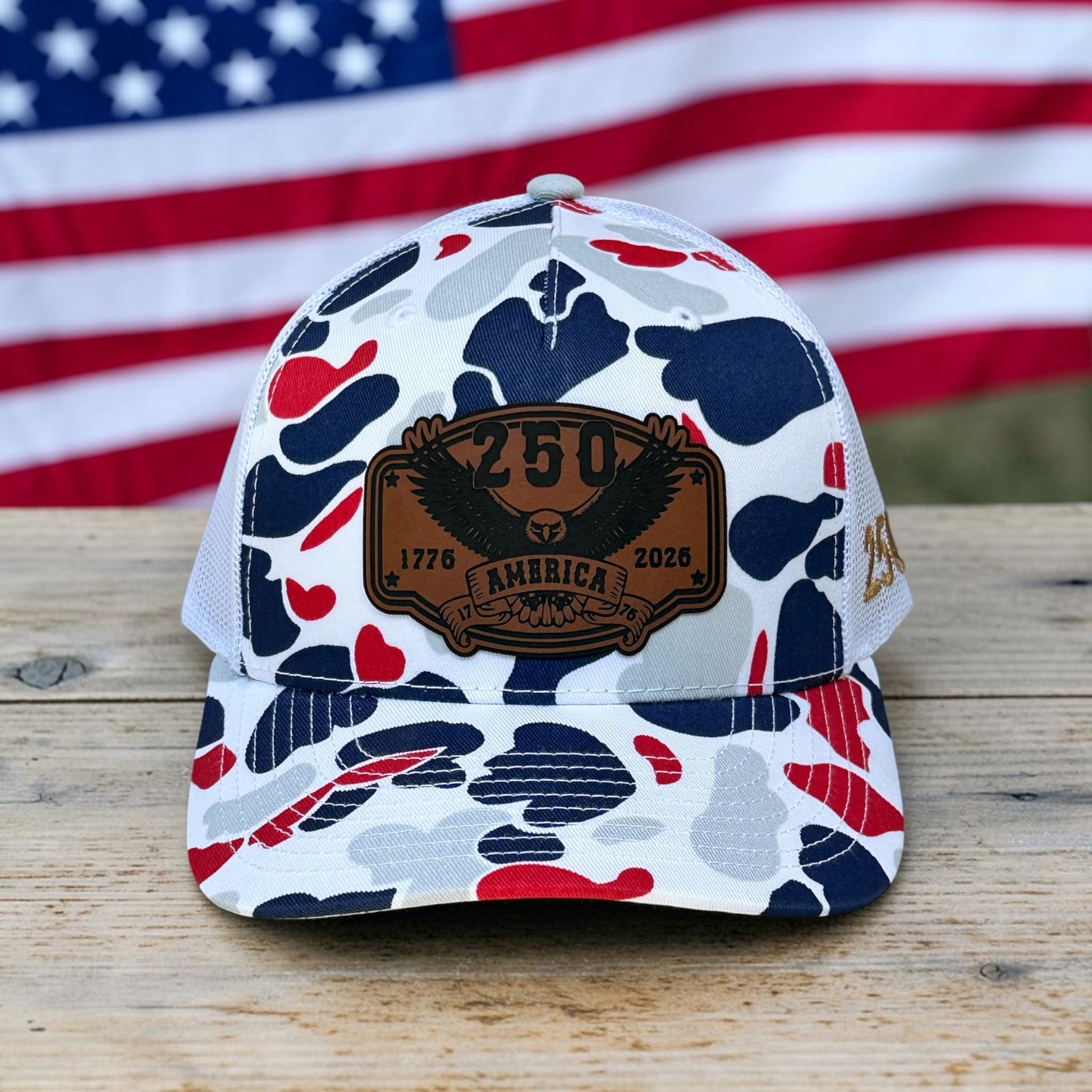 Red, white, and blue camouflage pattern hat with a leatherette patch that says "250 America, 1776-2026", on wooden surface with American flag in background