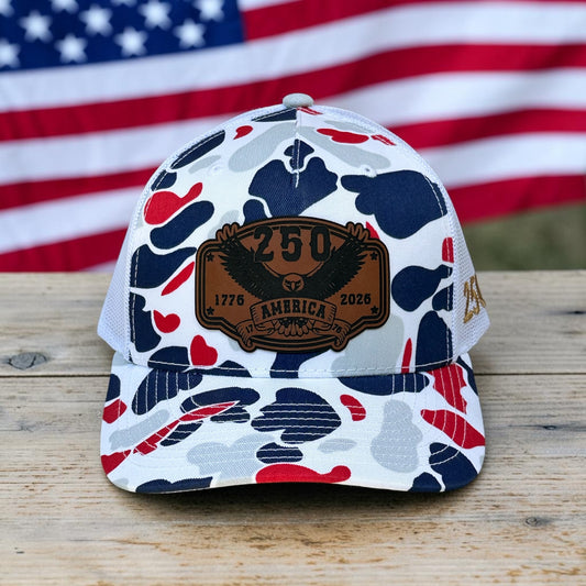 Red, white, and blue camouflage pattern hat with a leatherette patch that says "250 America, 1776-2026", on wooden surface with American flag in background