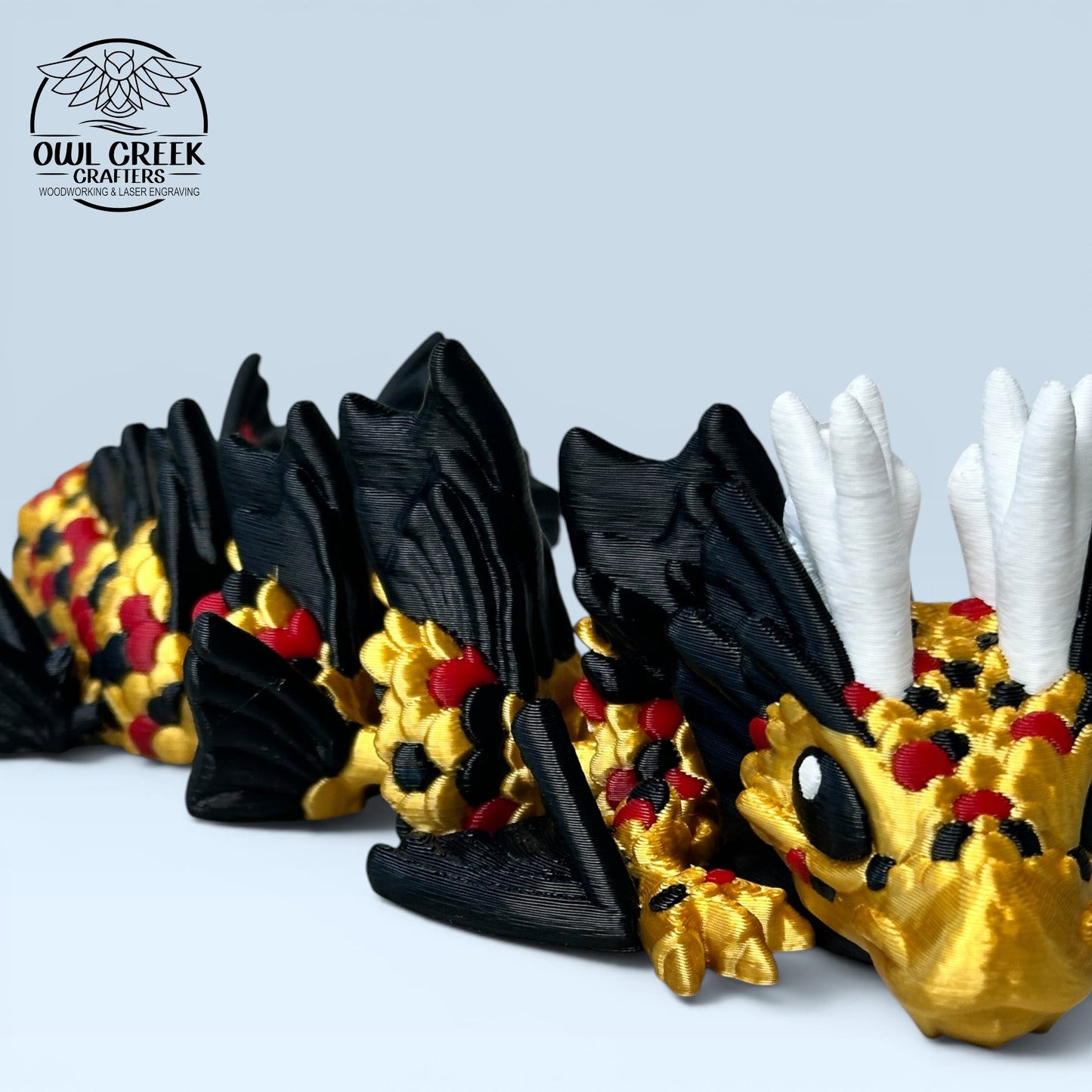 A 3D-printed articulated dragon figurine in black, gold, and white, posed on a light blue surface with the Owl Creek Crafters logo in the upper left.