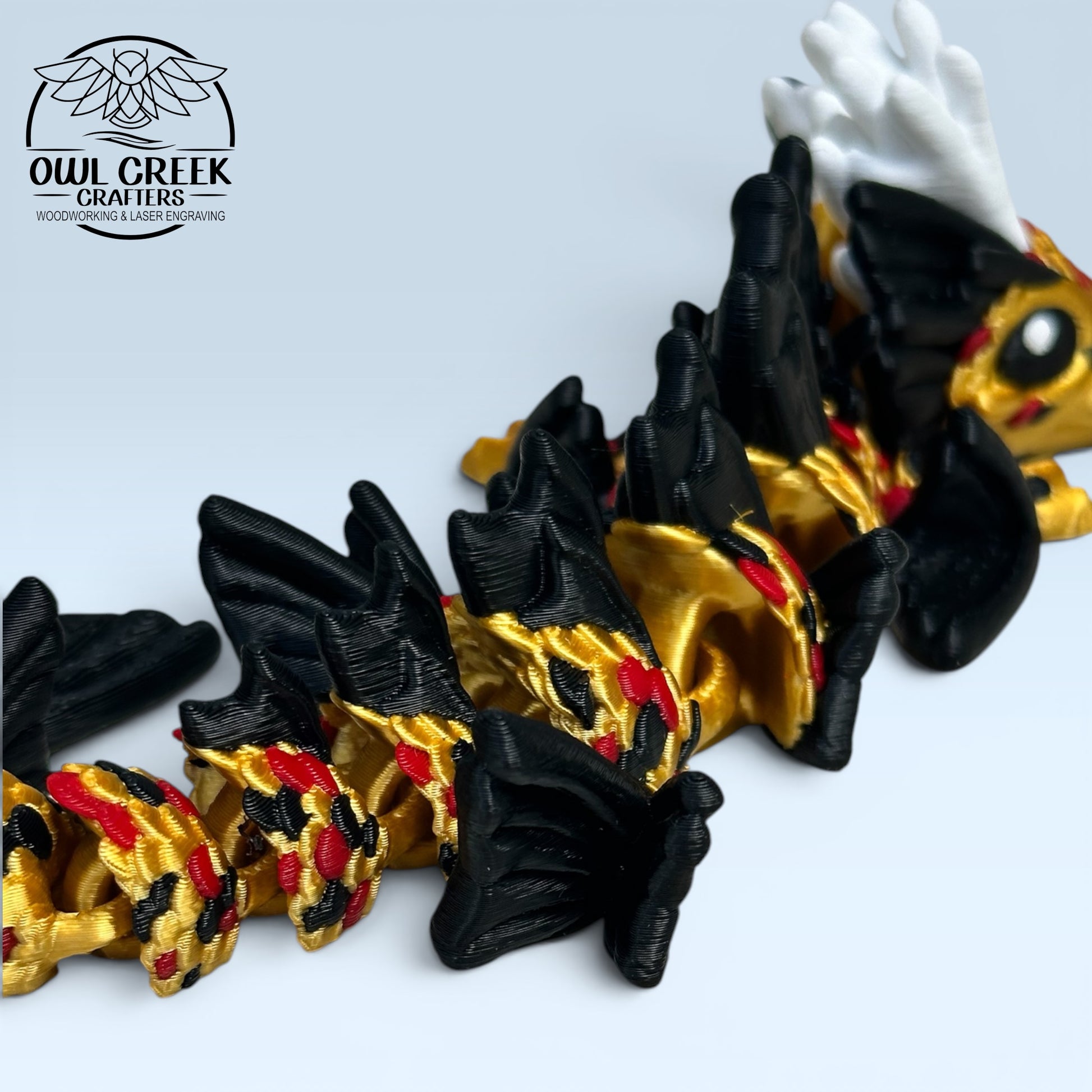 A 3D-printed articulated dragon figurine in black, gold, and white, posed on a light blue surface with the Owl Creek Crafters logo in the upper left.