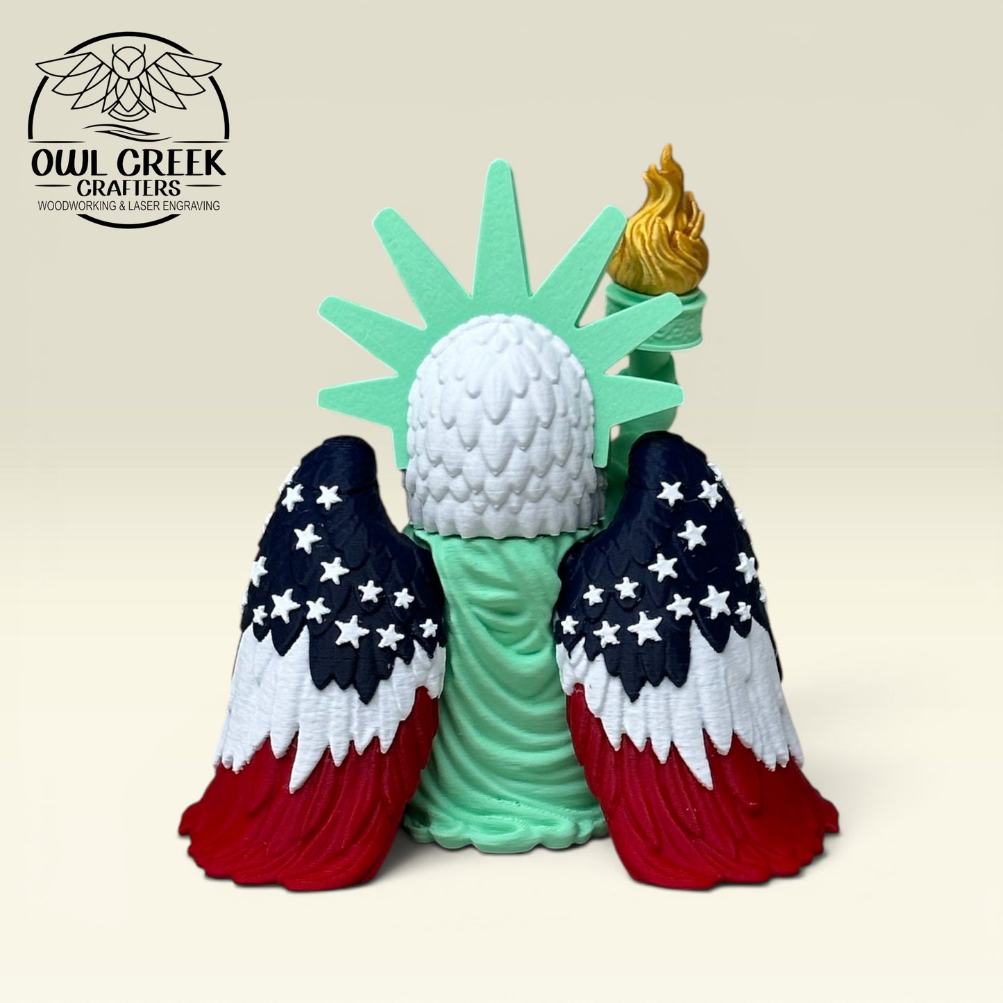 The back side of a bald eagle dressed as the Statue of Liberty with American flag wings, with a beige background and the Owl Creek Crafters Logo