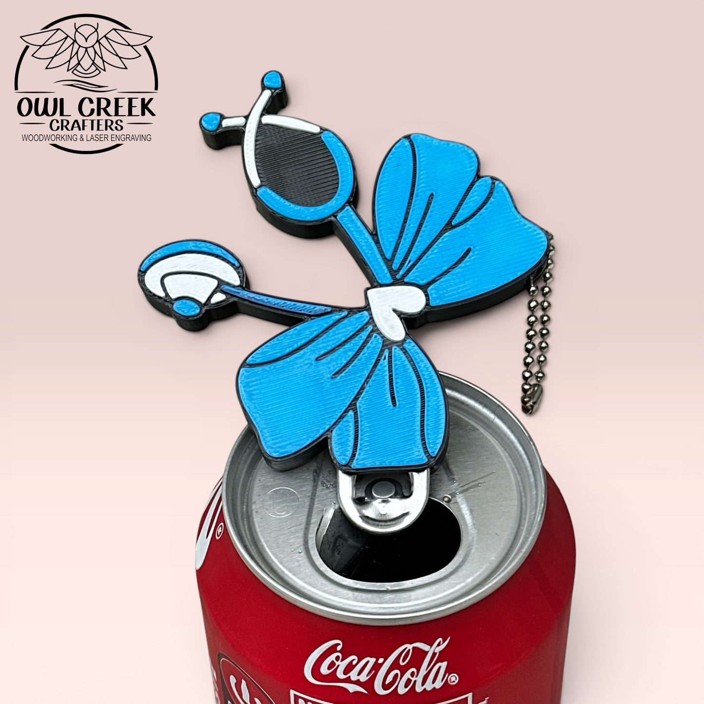 Blue stethescope bow bottle opener with chain on a Coca-Cola can, with a beige background and an Owl Creek Crafters logo