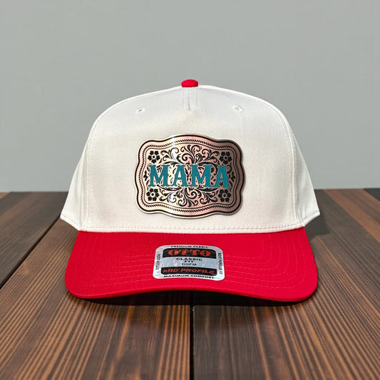 Two-tone cap with white top and red bill with a bronze acrylic patch with the text 'MAMA' in turquoise on a wooden surface with a grey background