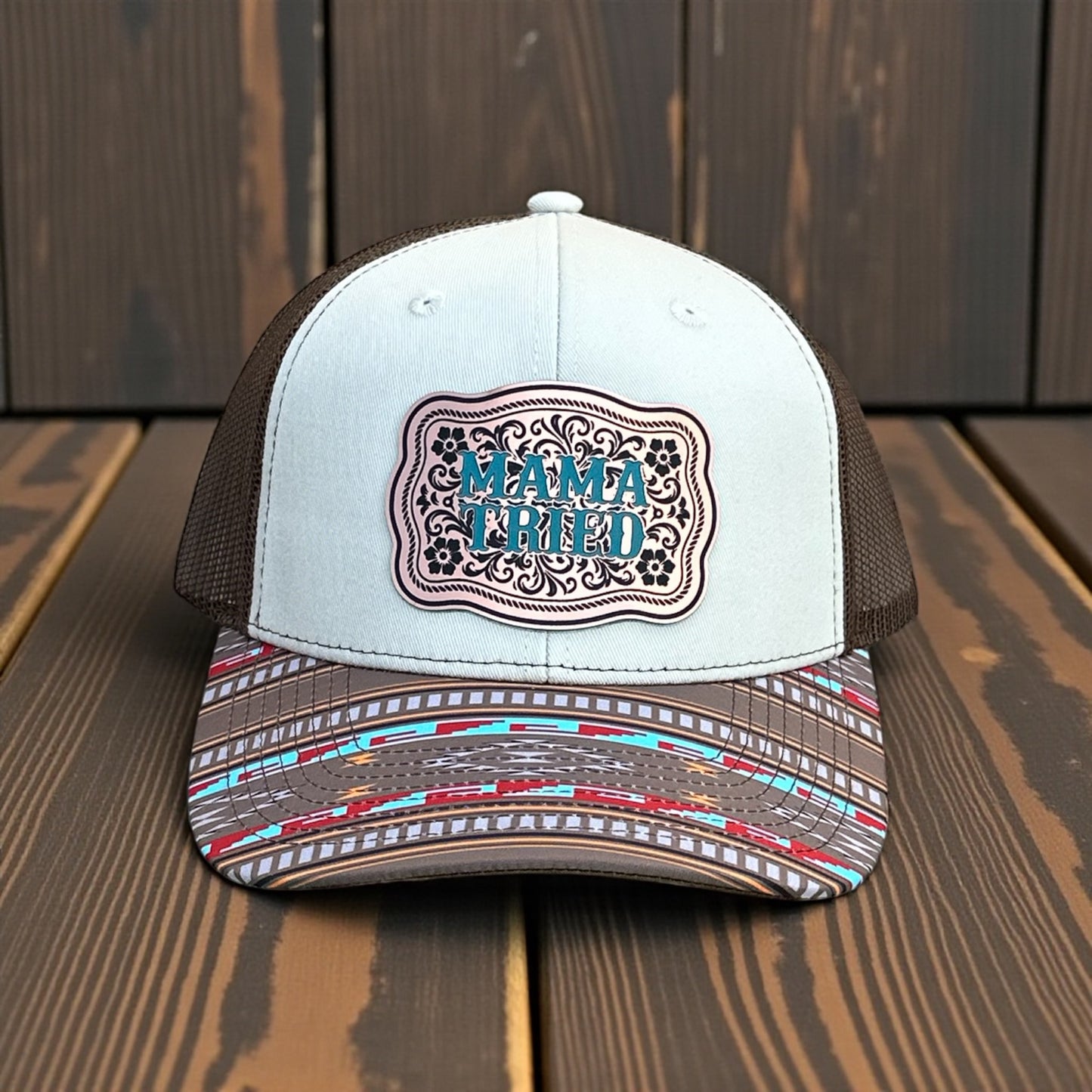 Tri-color trucker hat with a beige top, brown mesh back, and multi colored southwestern pattern bill with a bronze acrylic patch with the text 'Mama Tried' in turquoise, on a wooden surface and a wooden background