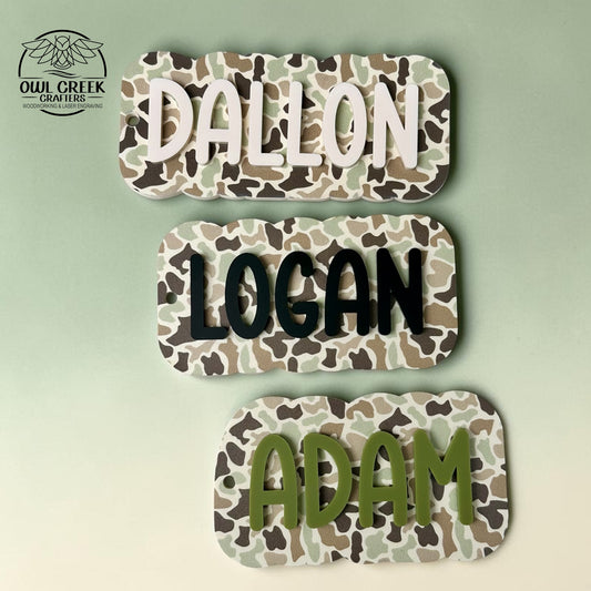 Three personalized name plaques with a camouflage pattern, displayed vertically on a light green background. The top plaque reads 'DALLON' in cream, the middle reads 'LOGAN' in black, and the bottom reads 'ADAM' in olive green. The Owl Creek Crafters logo is in the top left corner.