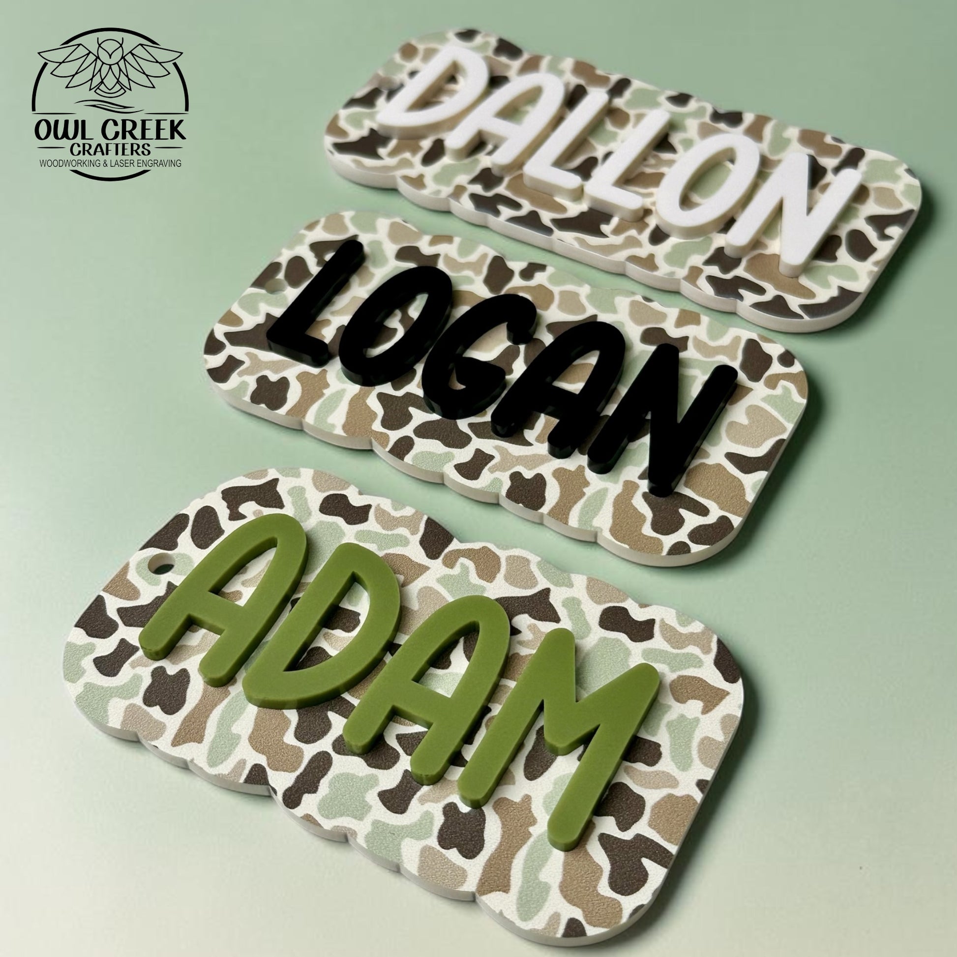 Three personalized name plaques with a camouflage pattern, featuring the names DALLON in cream, LOGAN in black, and ADAM in olive green, displayed on a light green background. The Owl Creek Crafters logo is in the top left corner.