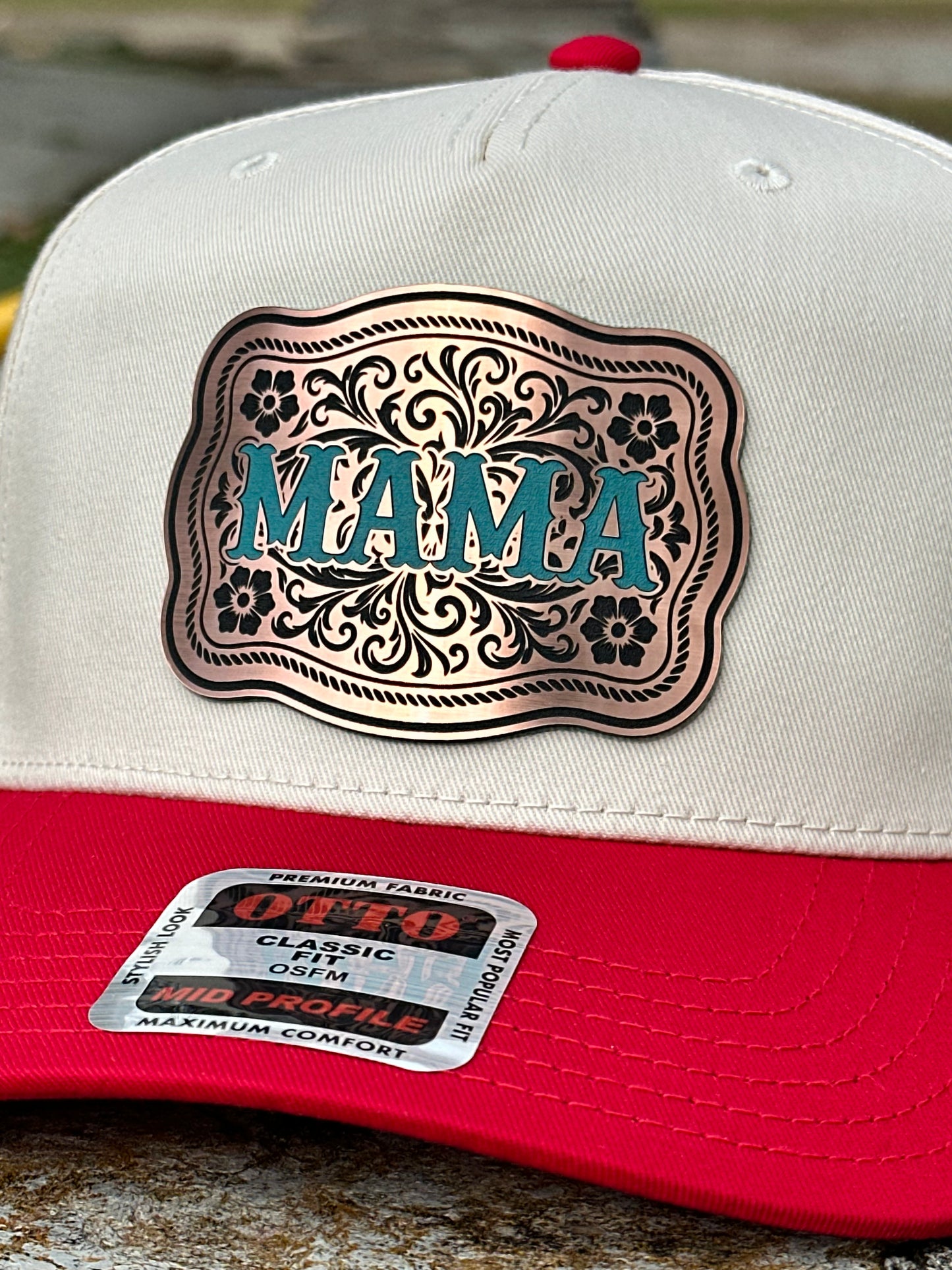 Close up of a two-tone cap with a white top and red bill and a bronze acrylic patch with the text 'MAMA' in turquoise on a textured background