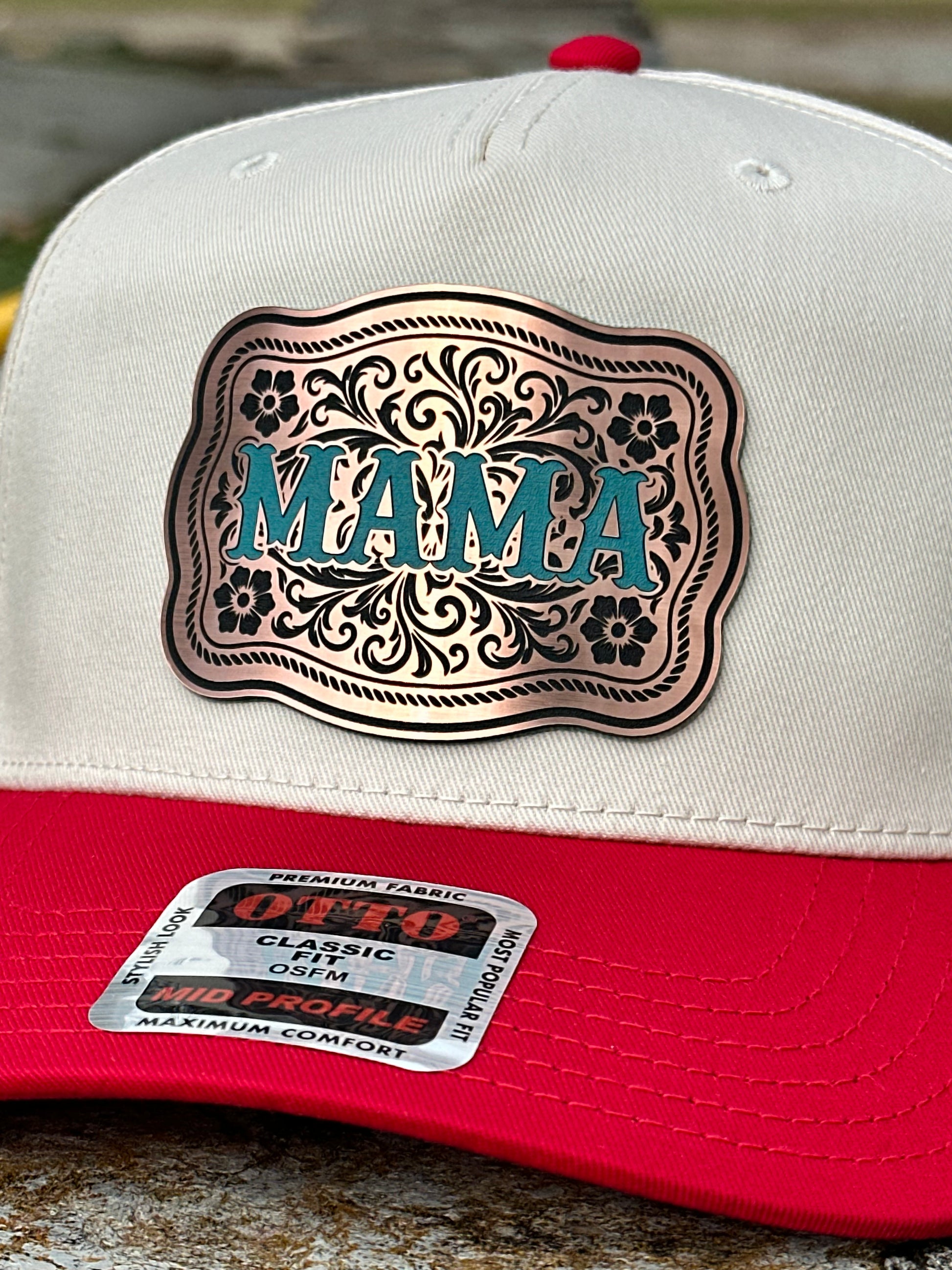 Close up of a two-tone cap with a white top and red bill and a bronze acrylic patch with the text 'MAMA' in turquoise on a textured background
