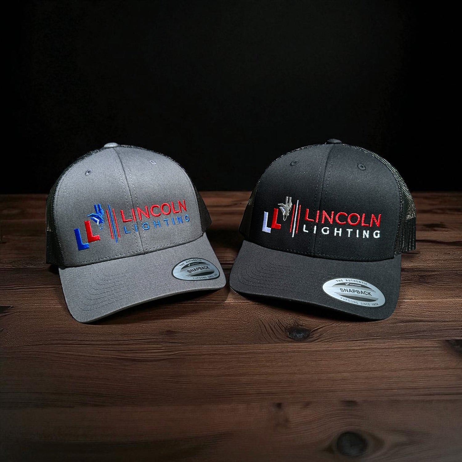 Two trucker caps, one gray one black, with en embroidered Lincoln Lighting brand logo on a wooden surface with a black background