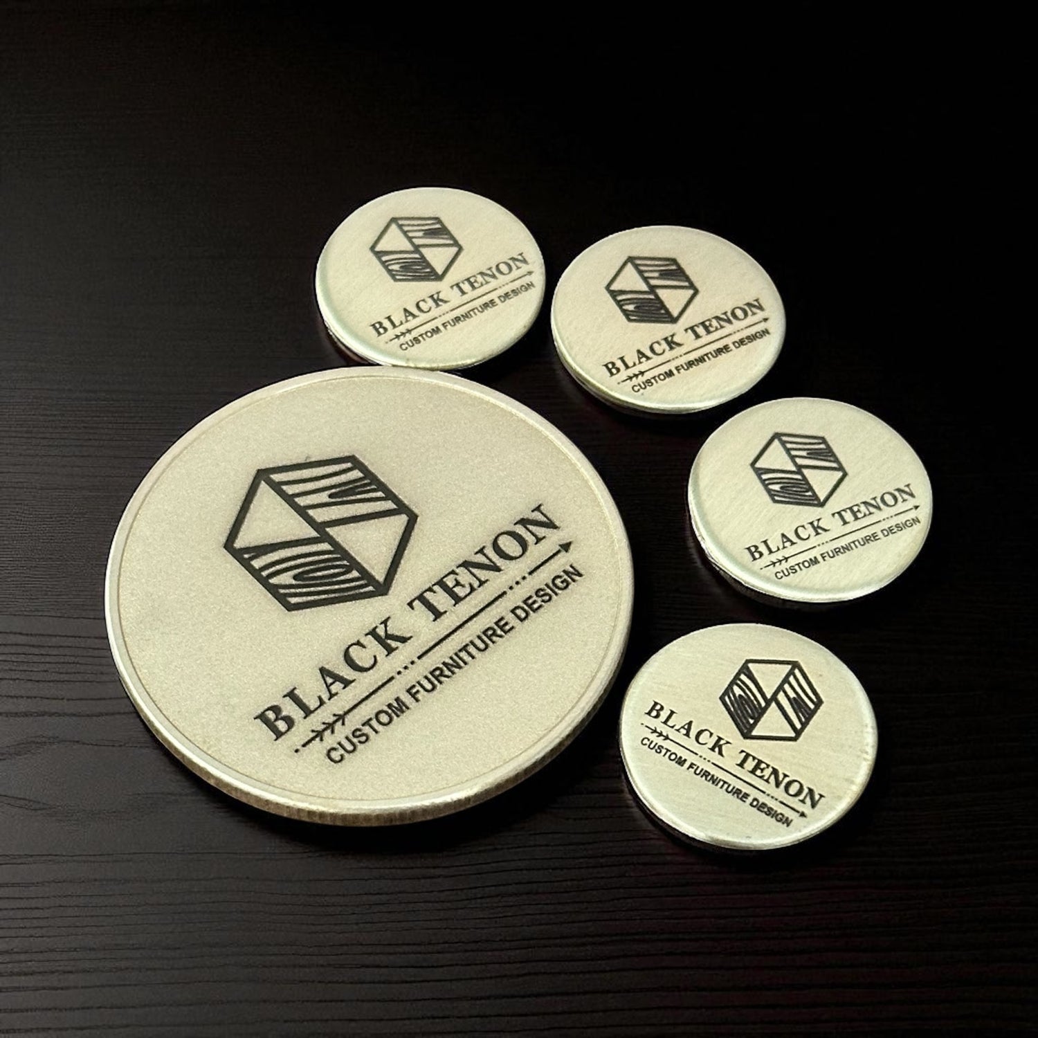 Custom laser engraved set of brass maker's mark coins with 'Black Tenon' branding on a dark surface