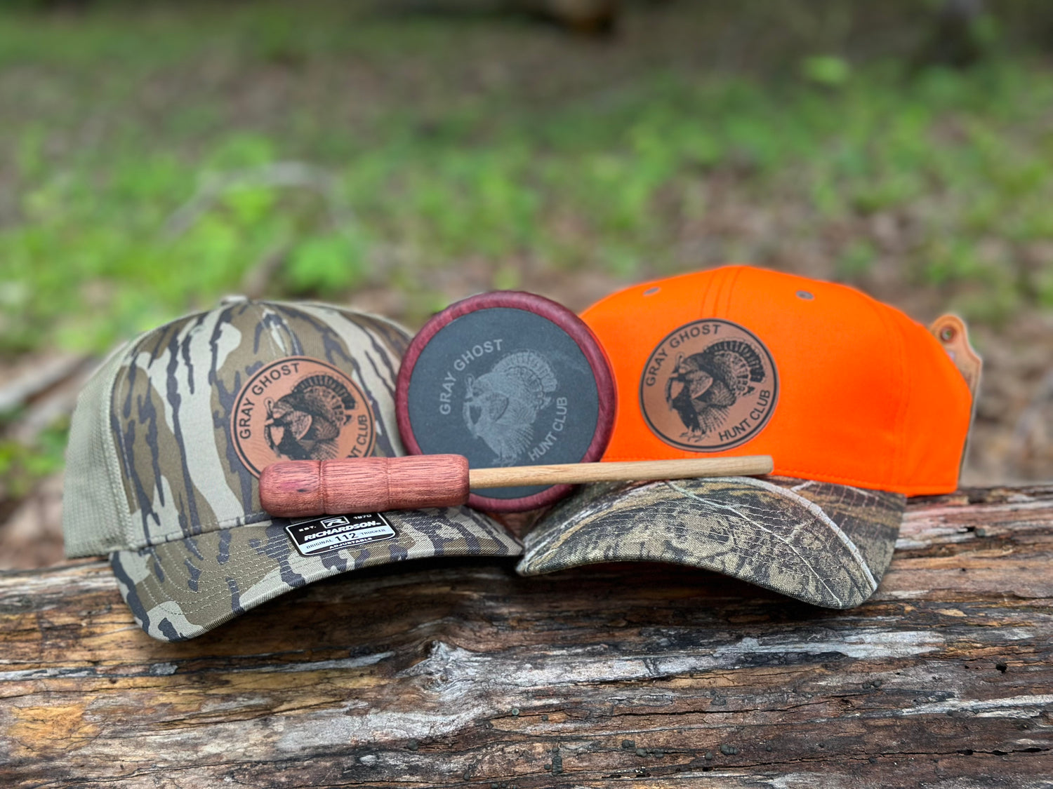 Two custom trucker hats with laser engraved leatherette patches with a turkey logo and a custom handmade turkey call. One hat is a green camoflage, the other is a two-tone hat with an orange top and camoflage bill. The hats are sitting on a log with woods in the background