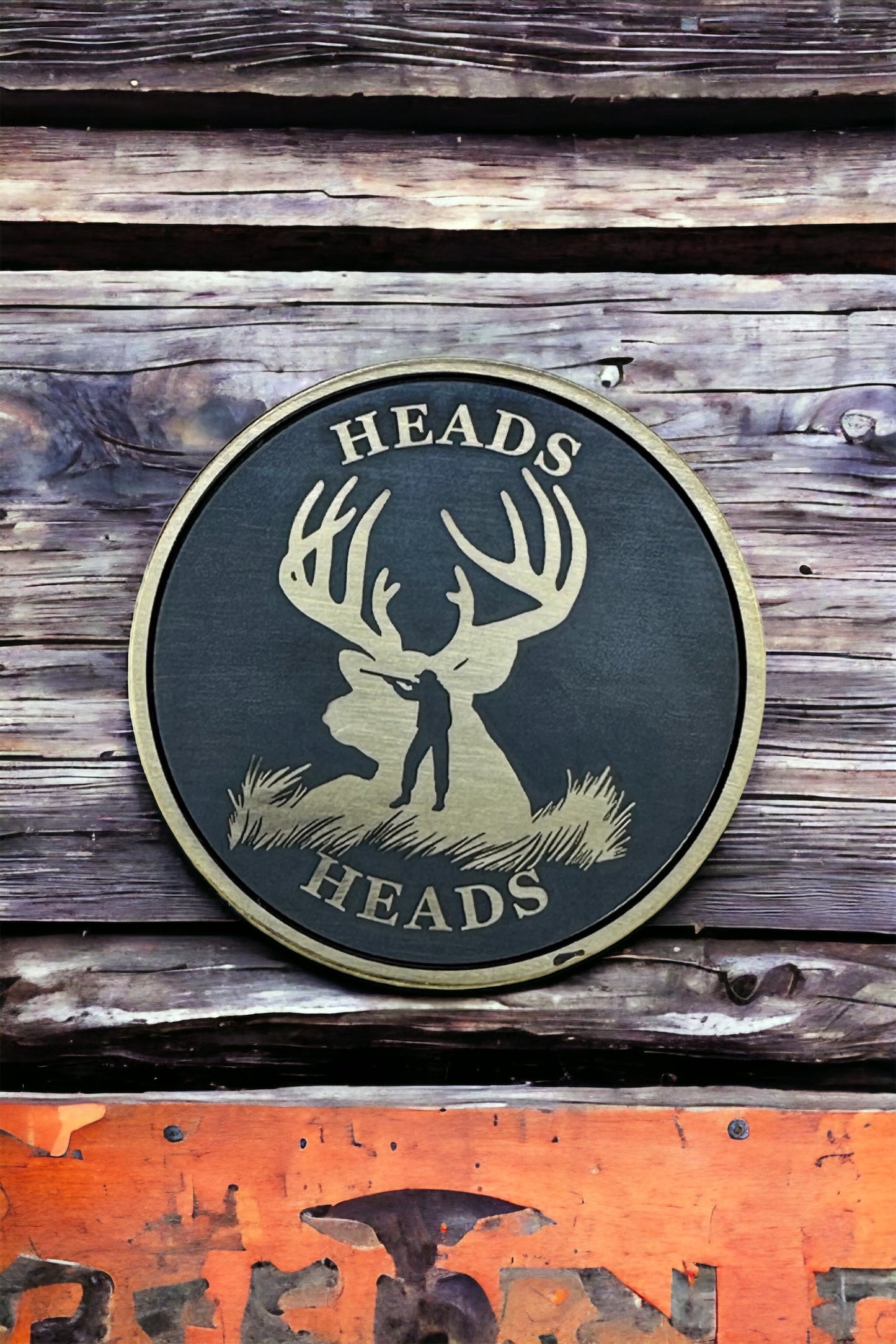 A double-sided solid brass coin with a laser engraved design of a deer and a hunter on it, with the text 'HEADS' around the edge. The coin is placed on a wooden surface.