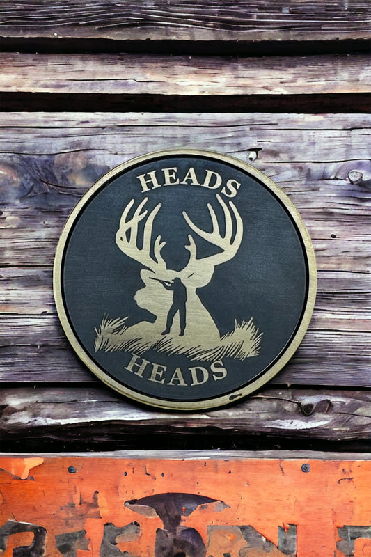 A double-sided solid brass coin with a laser engraved design of a deer and a hunter on it, with the text 'HEADS' around the edge. The coin is placed on a wooden surface.