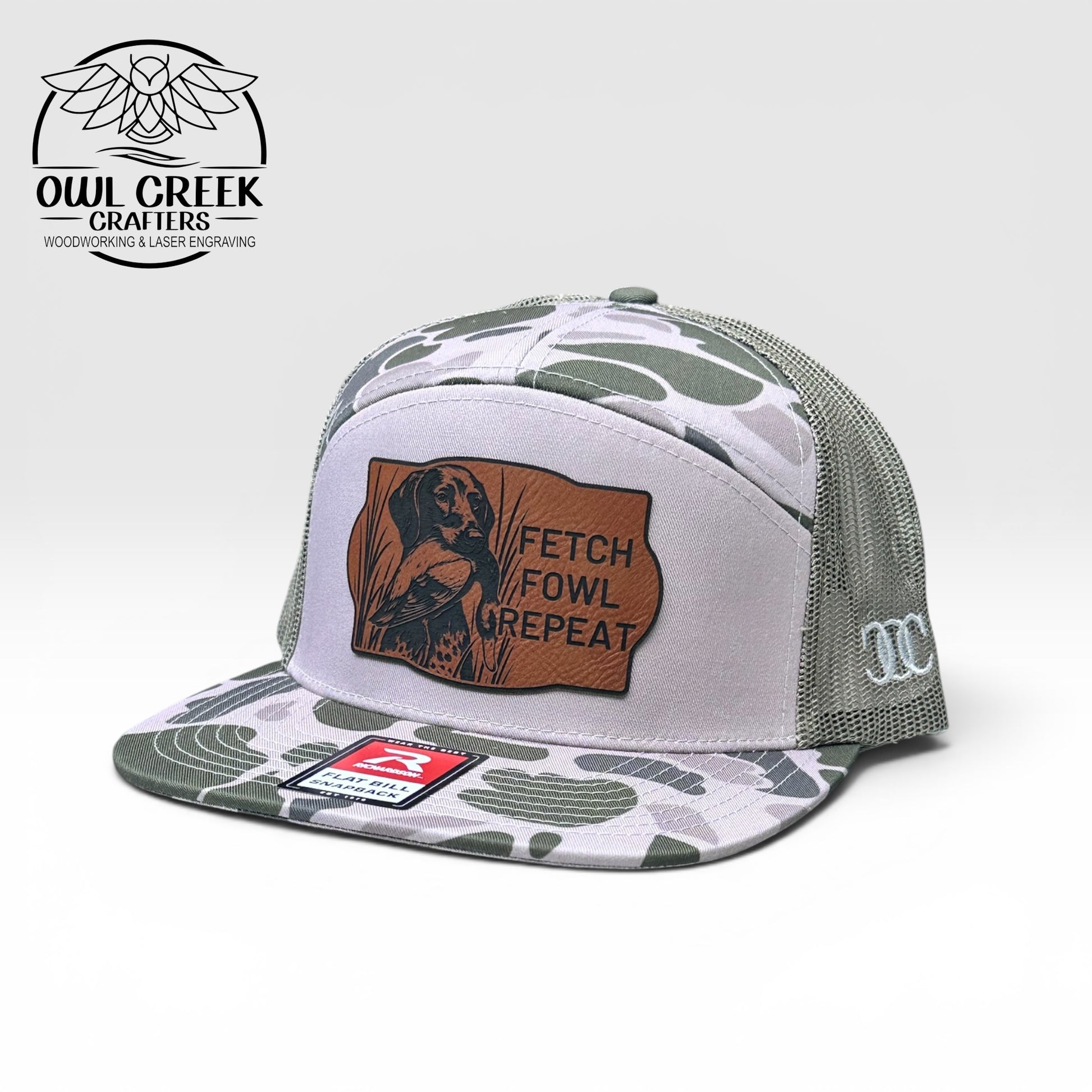 Trucker hat with green and grey camouflage pattern and leatherette patch on a white background, featuring 'Owl Creek Crafters' logo.