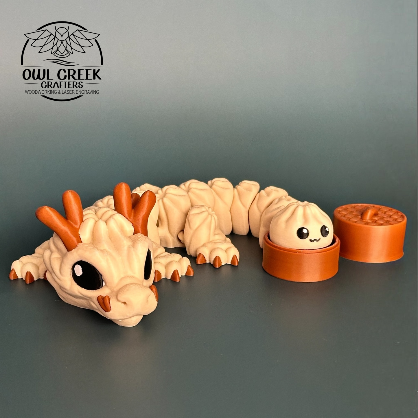 A 3d printed figurine of a cute, multi-segmented dumpling dragon in beige and brown, shown next to a small brown round container with a dumpling character and a lid. The background is a solid dark gray, and the Owl Creek Crafters logo is visible in the top left corner.
