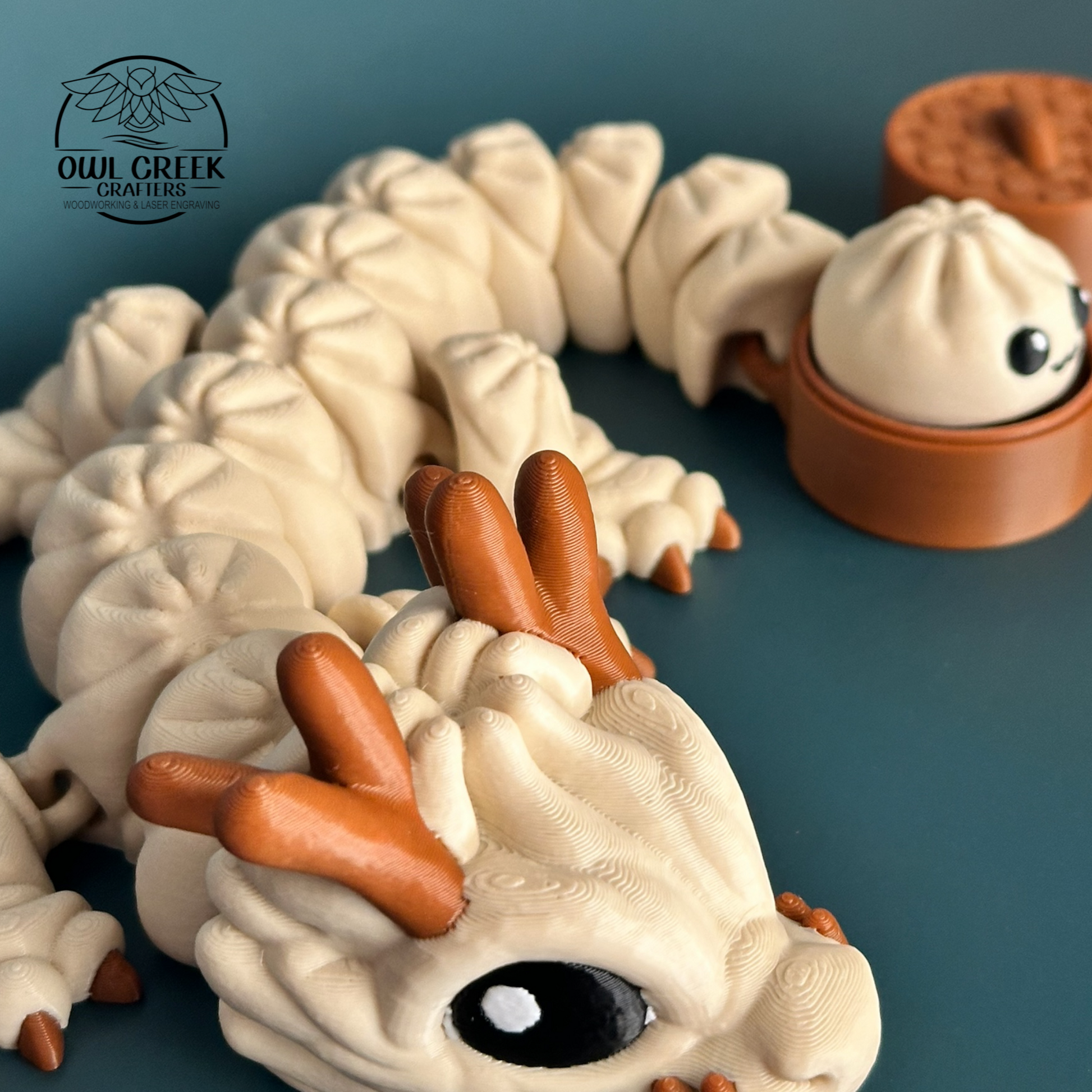 A close up of a 3d printed figurine of a cute, multi-segmented dumpling dragon in beige and brown, shown next to a small brown round container with a dumpling character and a lid. The background is a solid dark gray, and the Owl Creek Crafters logo is visible in the top left corner.