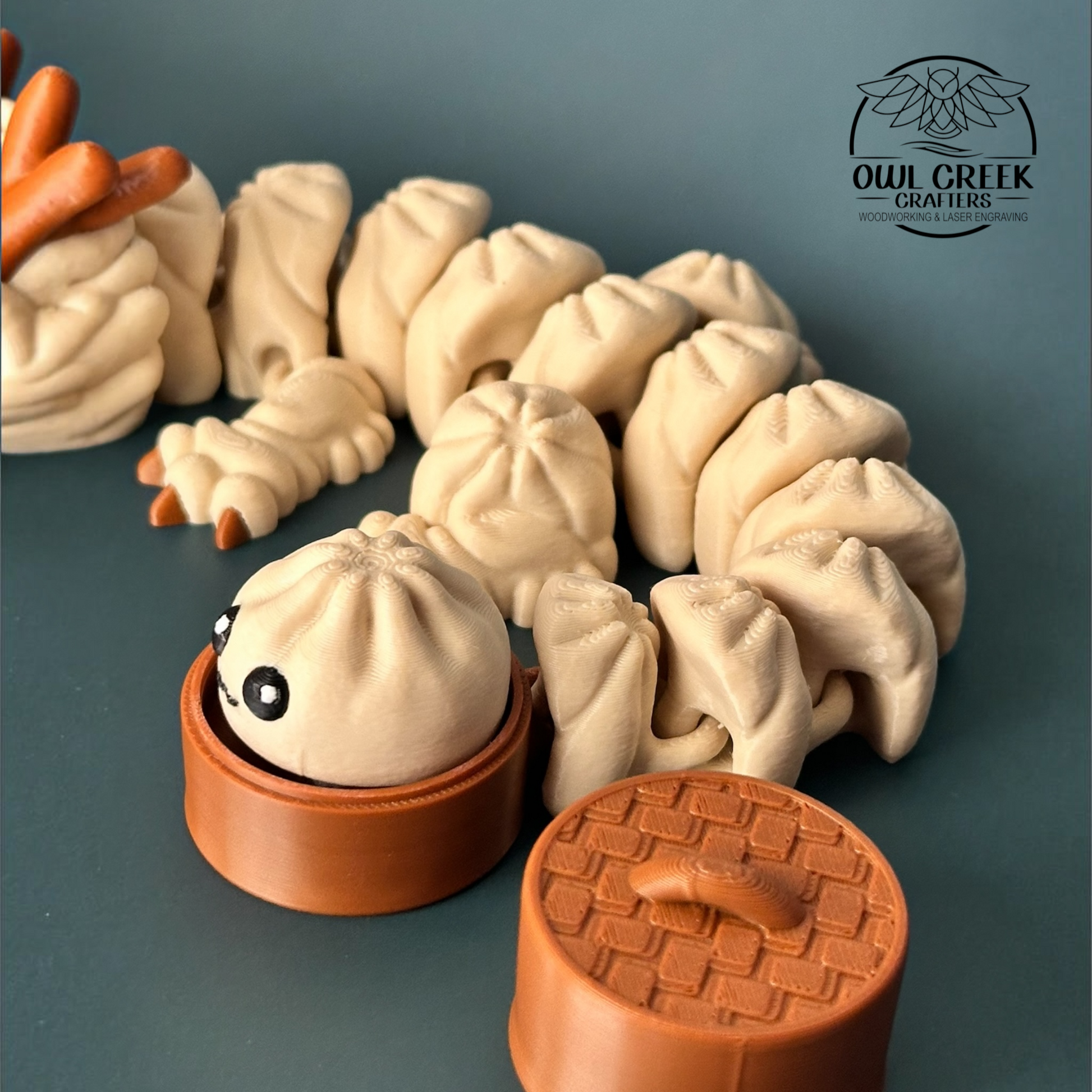 A close up 3d printed figurine of a cute, multi-segmented dumpling dragon in beige and brown, shown next to a small brown round container with a dumpling character and a lid. The background is a solid dark gray, and the Owl Creek Crafters logo is visible in the top right corner.
