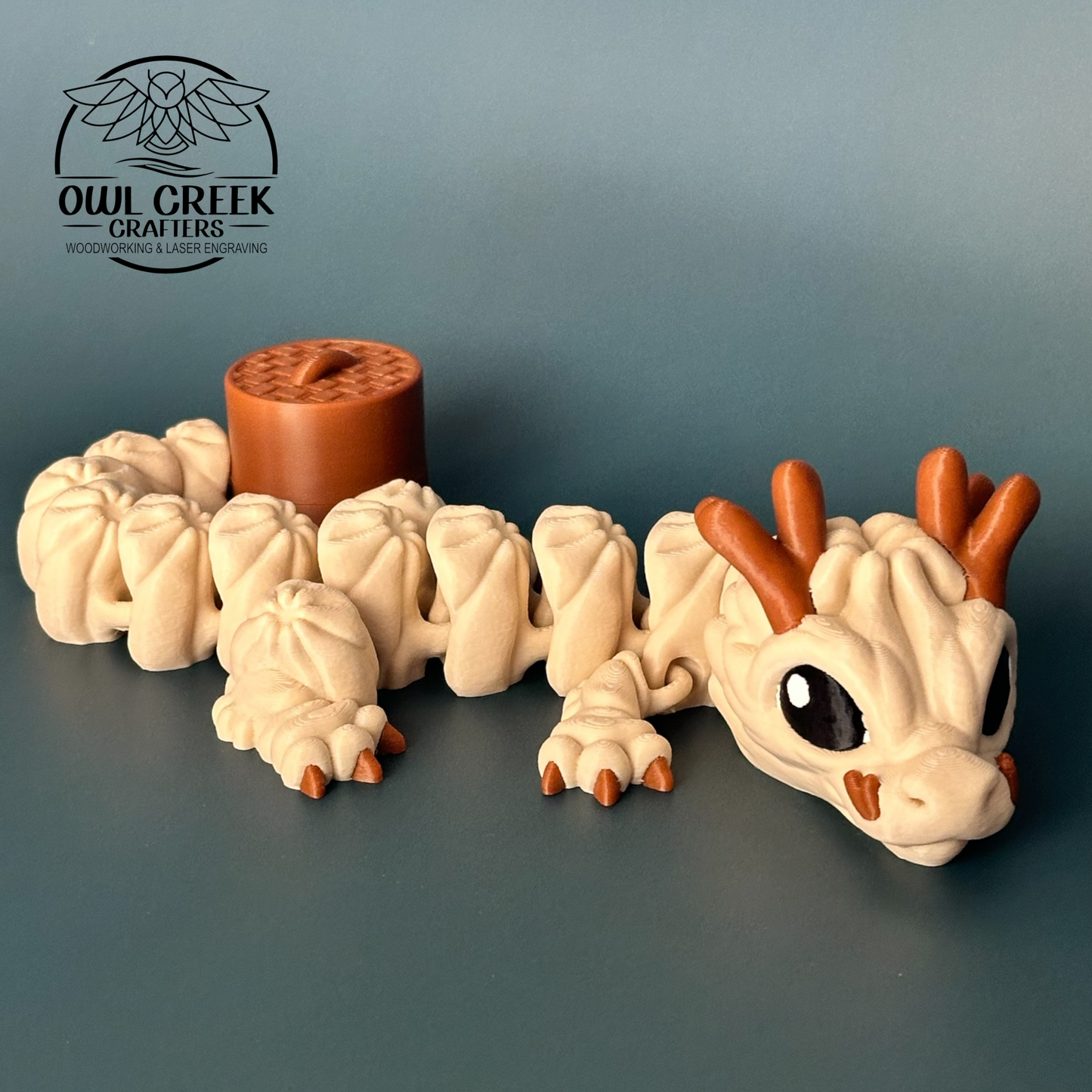 A 3d printed figurine of a cute, multi-segmented dumpling dragon in beige and brown, shown next to a small brown round container with a lid. The background is a solid dark gray, and the Owl Creek Crafters logo is visible in the top left corner.