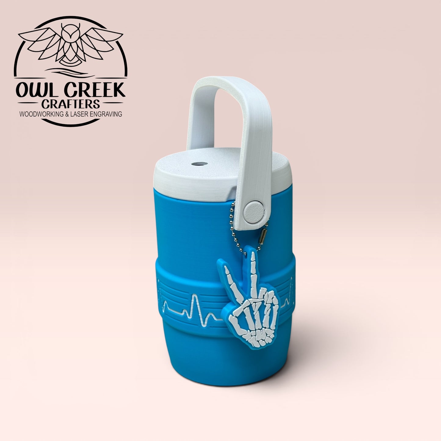 Blue can cooler with white skeleton hand keychain showing a peace sign design, with a beige background, featuring Owl Creek Crafters logo.