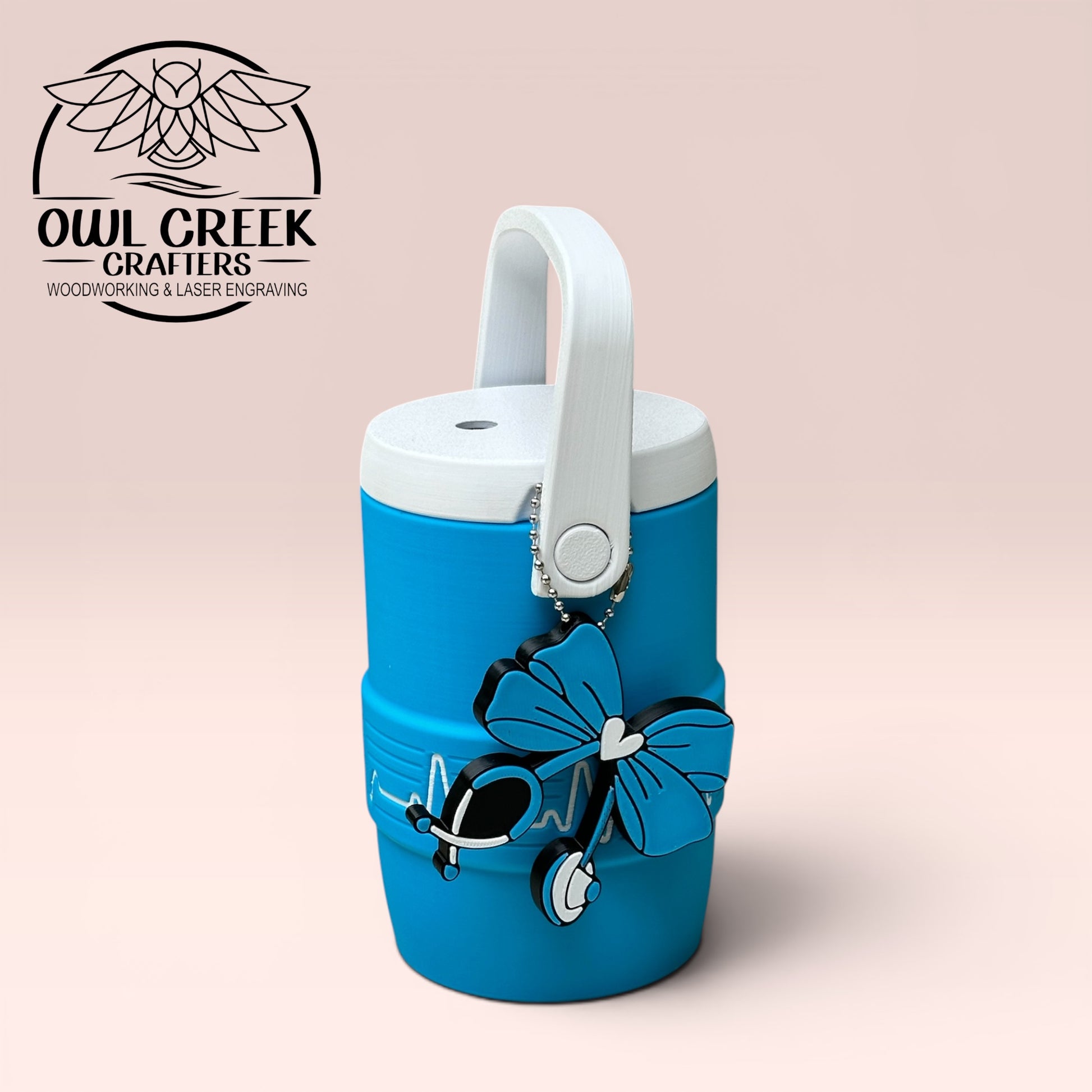 Blue can cooler coozie with a white EKG design, with a blue and white stethoscope bow design keychain, and white handle and lid on a beige background, featuring Owl Creek Crafters logo