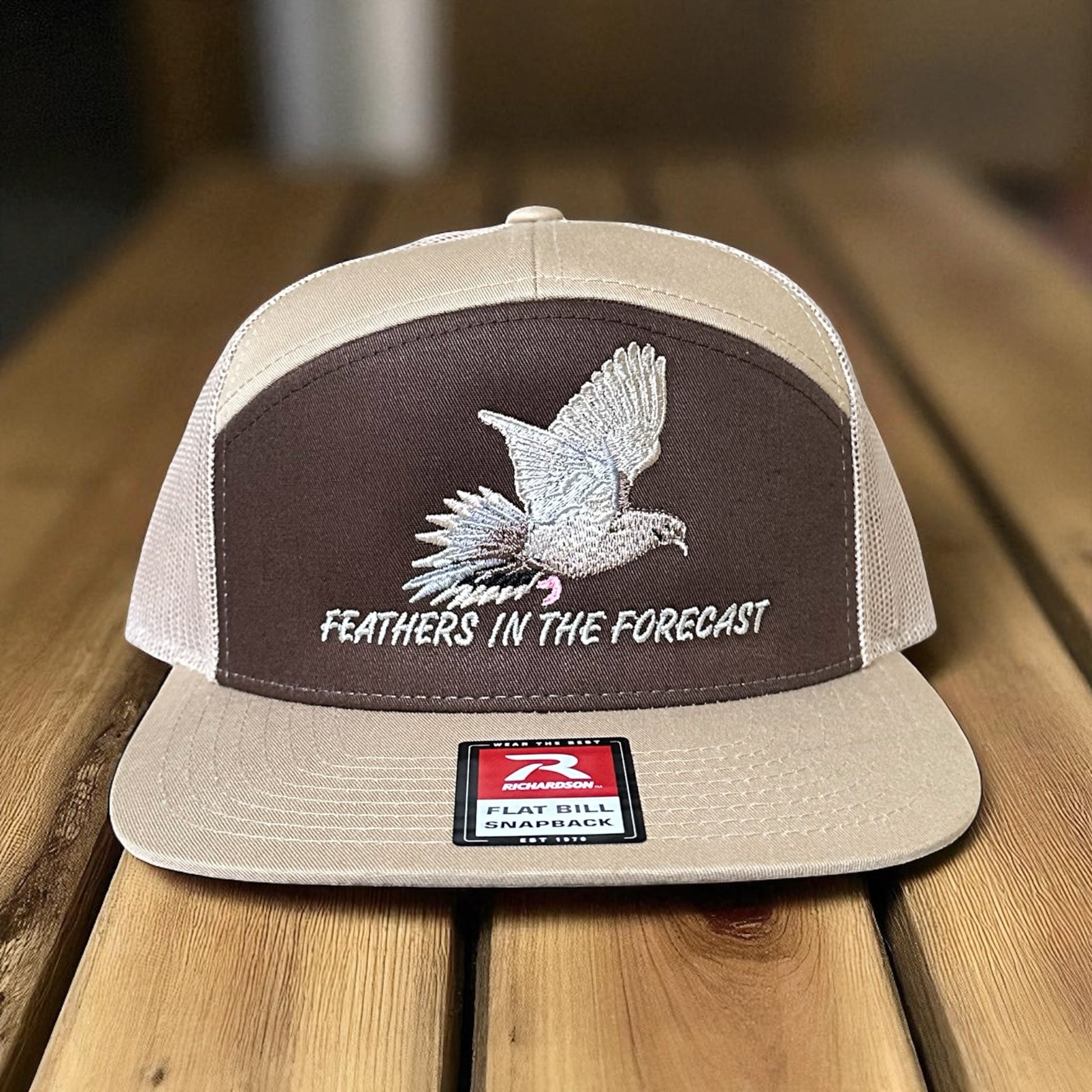 Brown and khaki trucker hat with an embroidered dove design and the text 'Feathers in the Forecast' on a wooden surface with a visible brand logo label