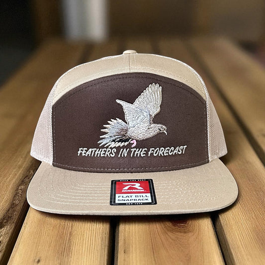 Brown and khaki trucker hat with an embroidered dove design and the text 'Feathers in the Forecast' on a wooden surface with a visible brand logo label