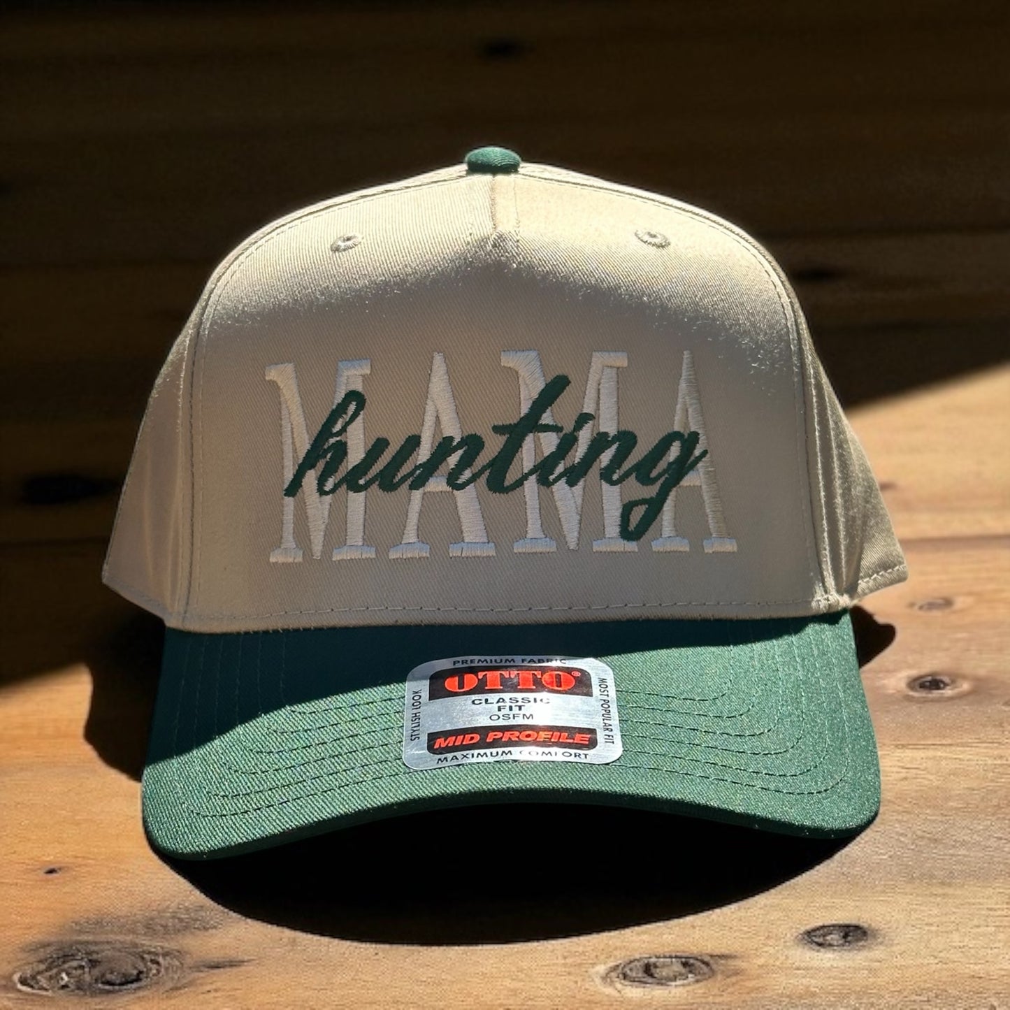 Two-tone trucker hat with beige top and green bill with embroidered text 'Mama hunting' , and a visible brand logo label on a wooden surface