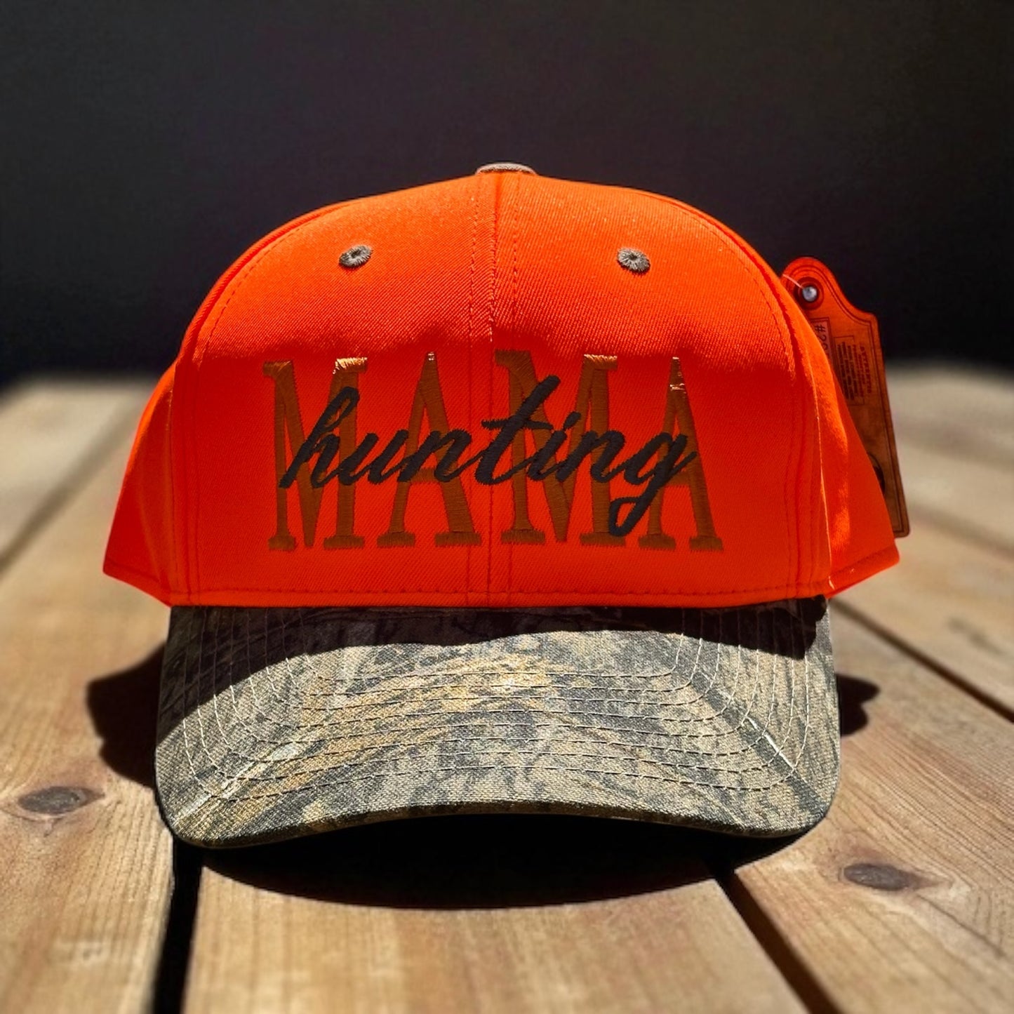 Two-tone trucker hat with orange top and camoflauge bill with embroidered text 'Mama hunting' on a wooden surface with a dark background