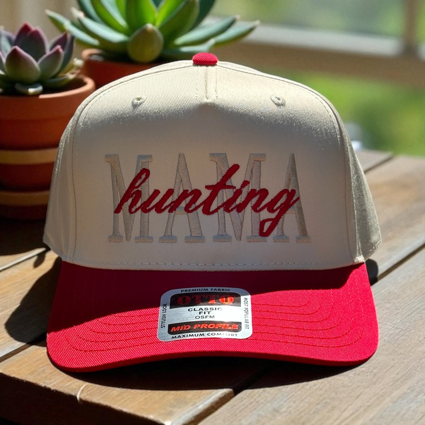 Two-tone trucker hat with beige top and red bill with embroidered text 'Mama hunting' , and a visible brand logo label on a wooden surface with plants in the background