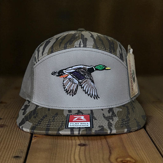 Camouflage and green trucker cap with an embroidered mallard duck design on a wooden surface and brown background