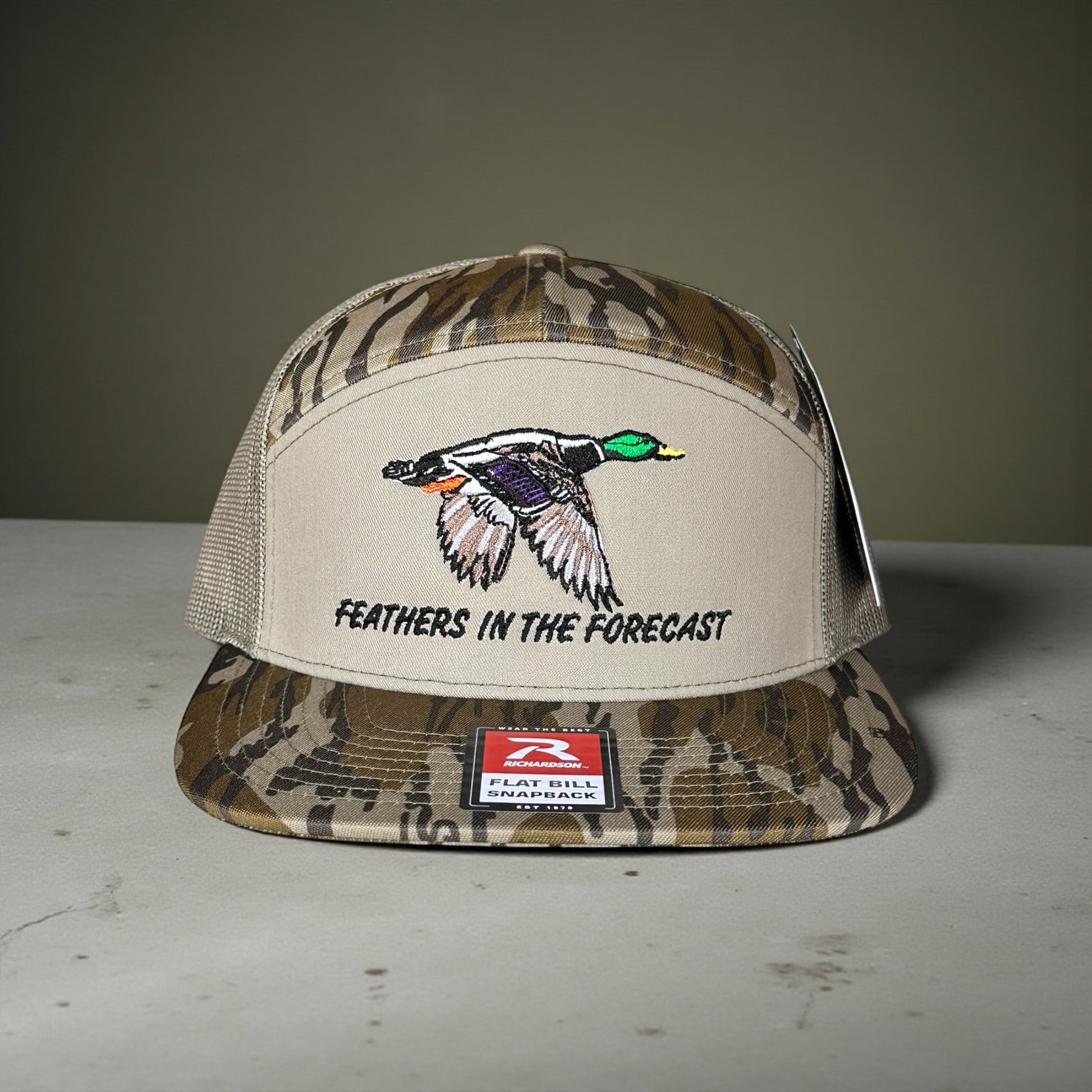 Camouflage and beige cap with an embroidered flying duck design with Feathers in the Forecast text on a gray surface and green background