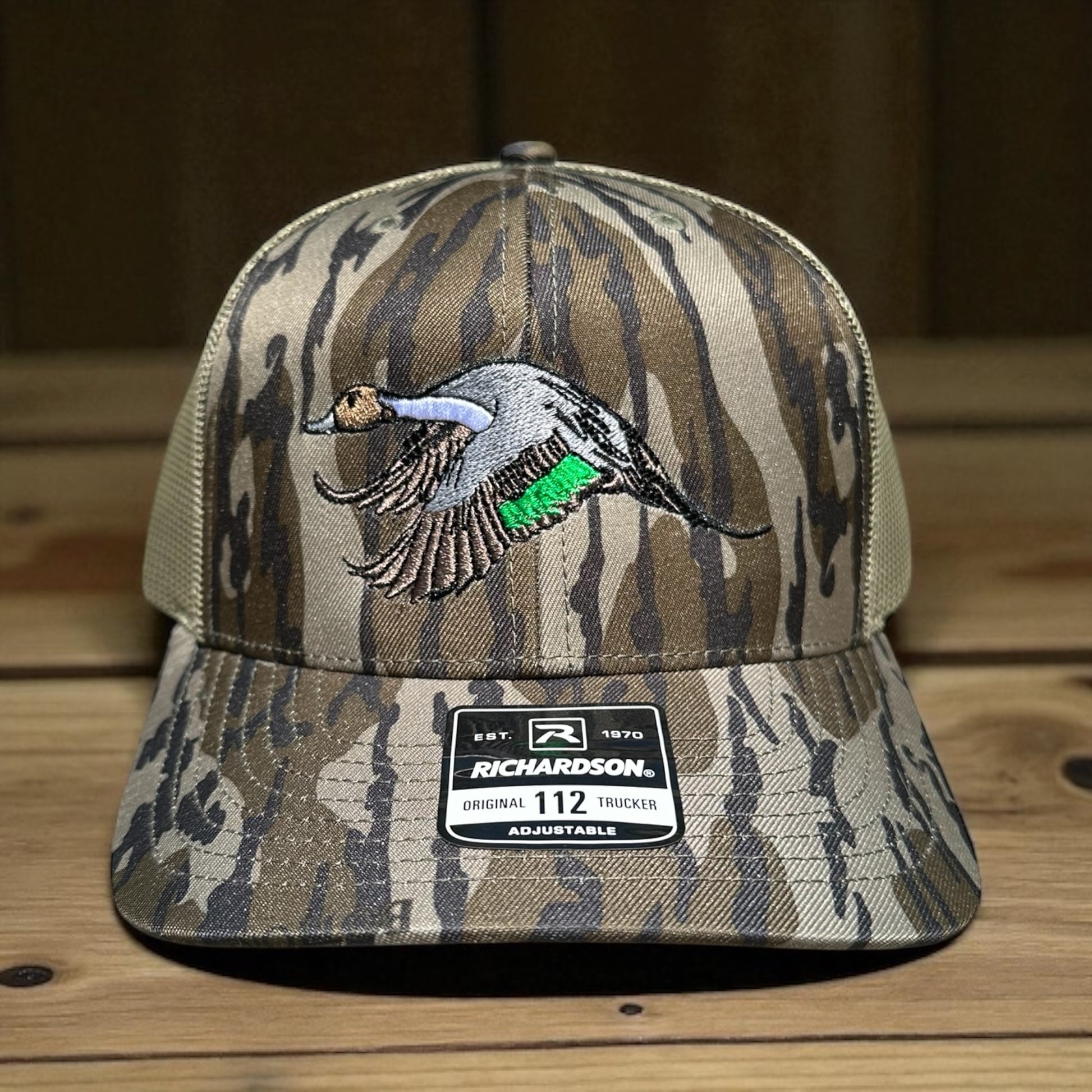 Camouflage trucker cap with an embroidered pintail duck design and a visible brand logo label on a wooden surface