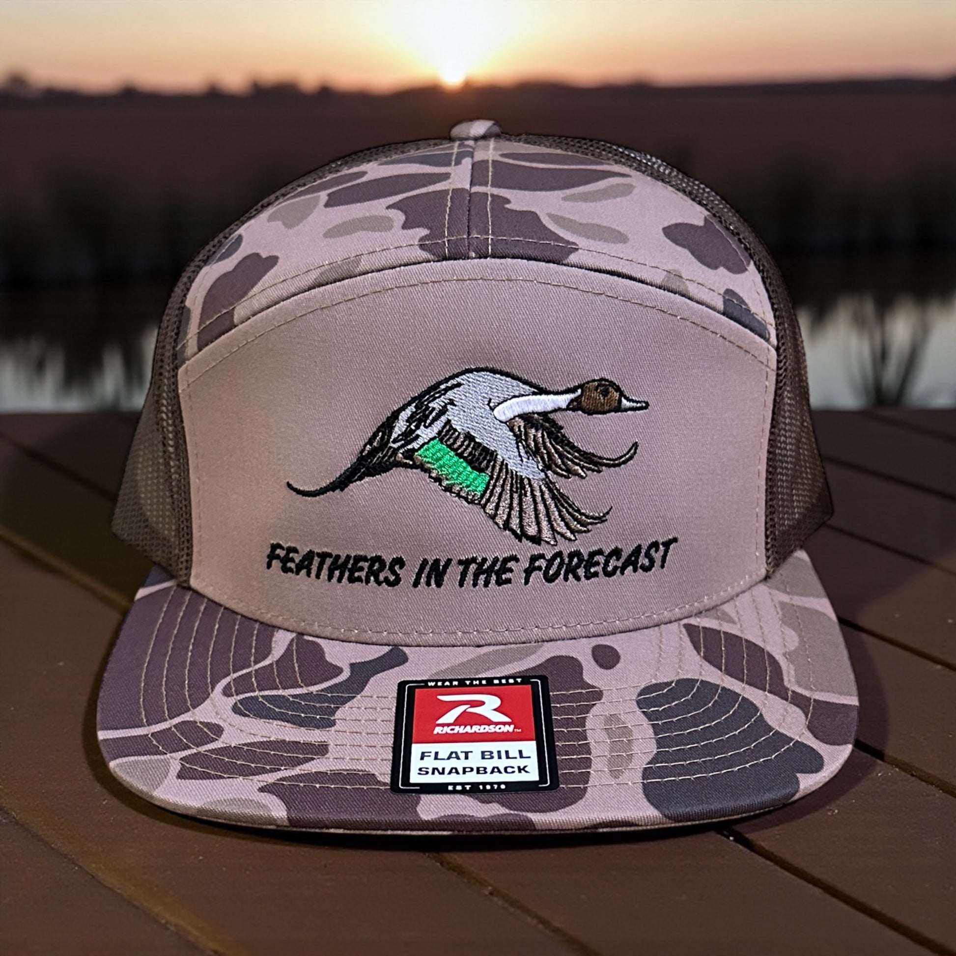 Camouflage trucker cap with an embroidered pintail duck design and text 'Feathers in the Forecast', with a visible brand logo label on a wooden surface with sunset lake background.