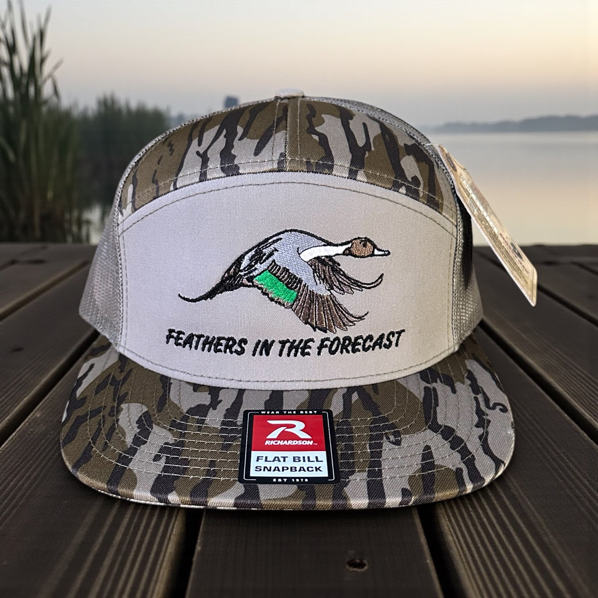 Camouflage trucker cap with and embroidered pintail duck design and text 'Feathers in the Forecast', and a visible brand logo label on a wooden surface with a lake background.